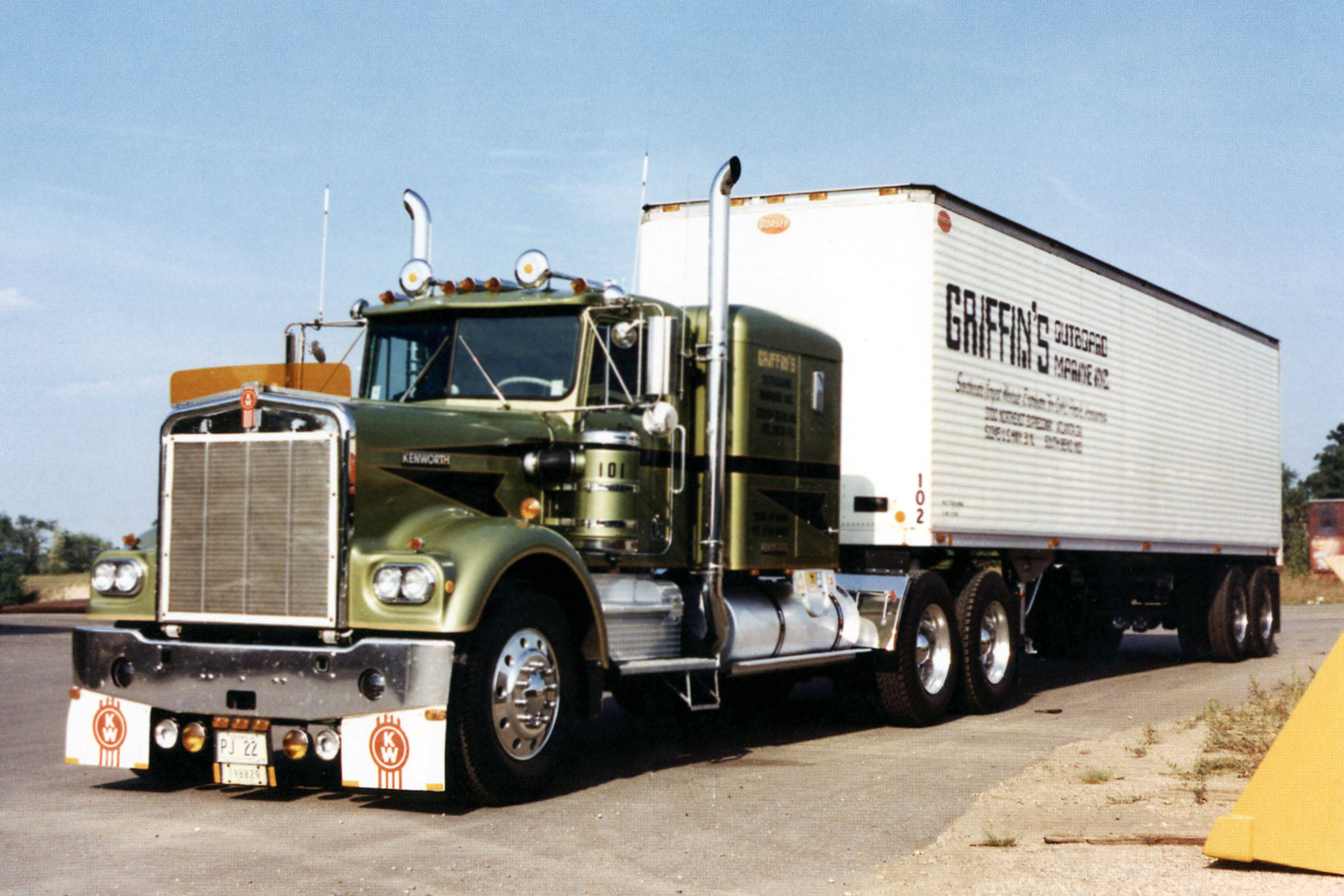Kenworth W900 桌布 壁紙 — 一輛復古橄欖綠色的Kenworth半掛卡車，帶有鍍鉻裝飾和白色拖車，拖車上標有'Griffin's Catering'，停在瀝青路面上，天空清澈湛藍。