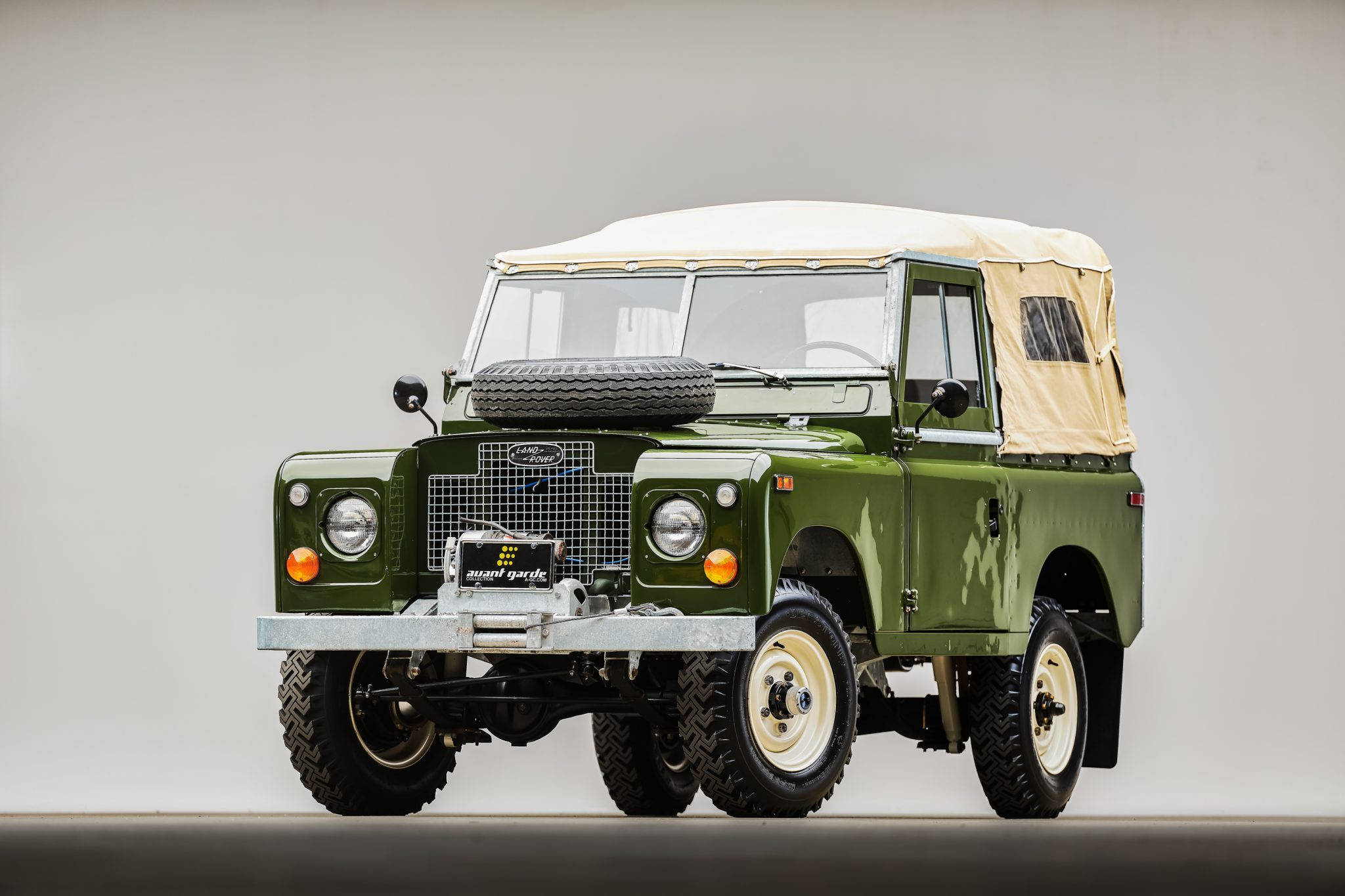 land rover iPhone 桌布 壁紙 — 一輛復古橄欖綠色的Land Rover，配備米色帆布車頂和前格柵上安裝的備用輪胎，在中性工作室背景前靜立。