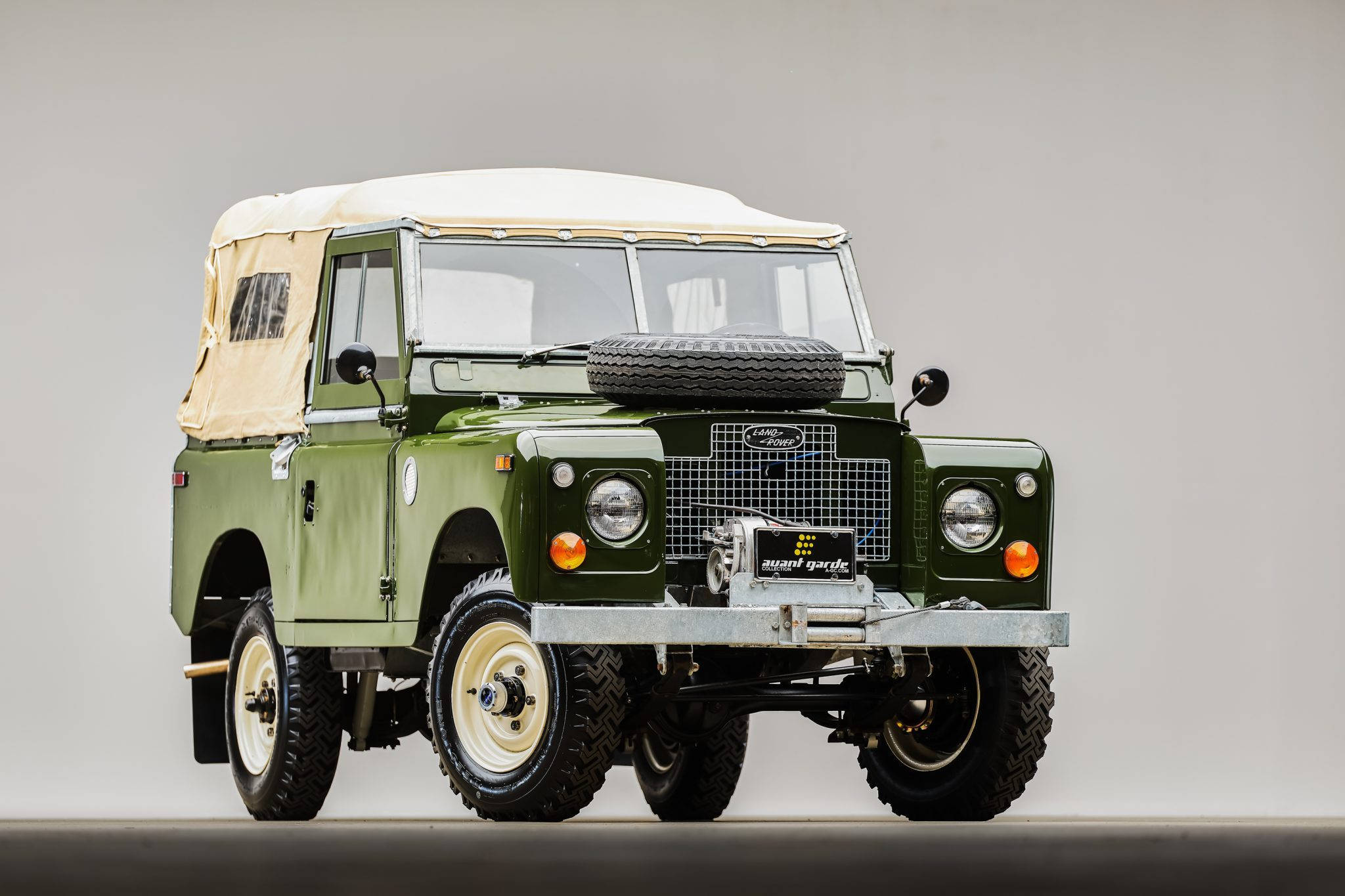 land rover iPhone 桌布 壁紙 — 一輛復古橄欖綠色的Land Rover，配有米色帆布車頂和安裝在引擎蓋上的備用輪胎，在中性工作室背景下蓄勢待發。