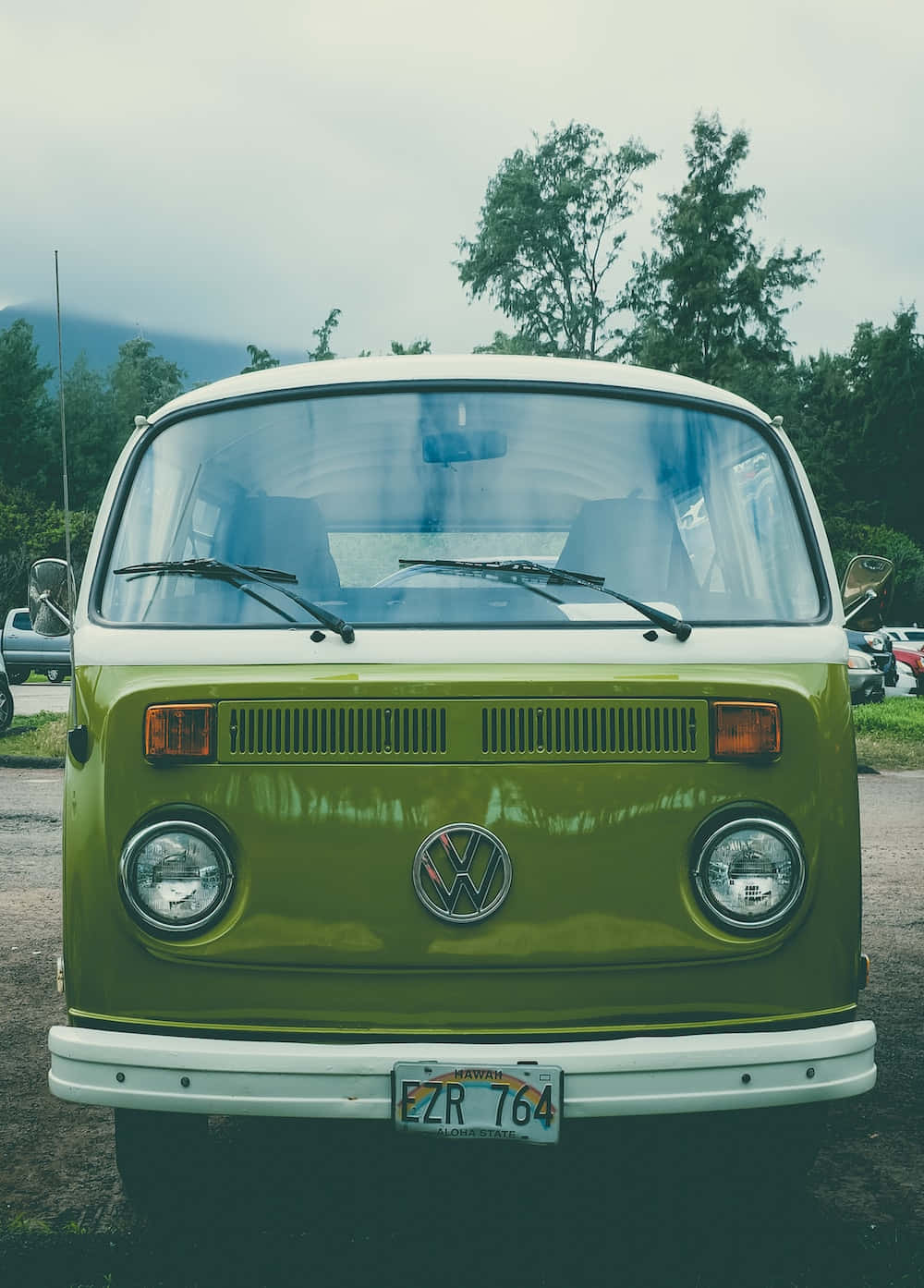 復古美學 桌布 壁紙 — 一輛復古橄欖綠色的福斯廂型車，帶有白色飾條，在陰天背景下，背景有樹木和山脈，車頭朝前。