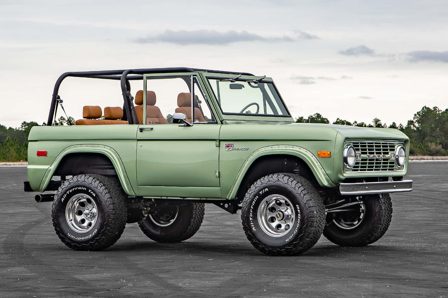 福特野馬越野車 桌布 壁紙 — 一輛復古橄欖綠色的福特 Bronco，配備米色內裝和超大輪胎，停在鋪砌的停車場上，天空多雲。