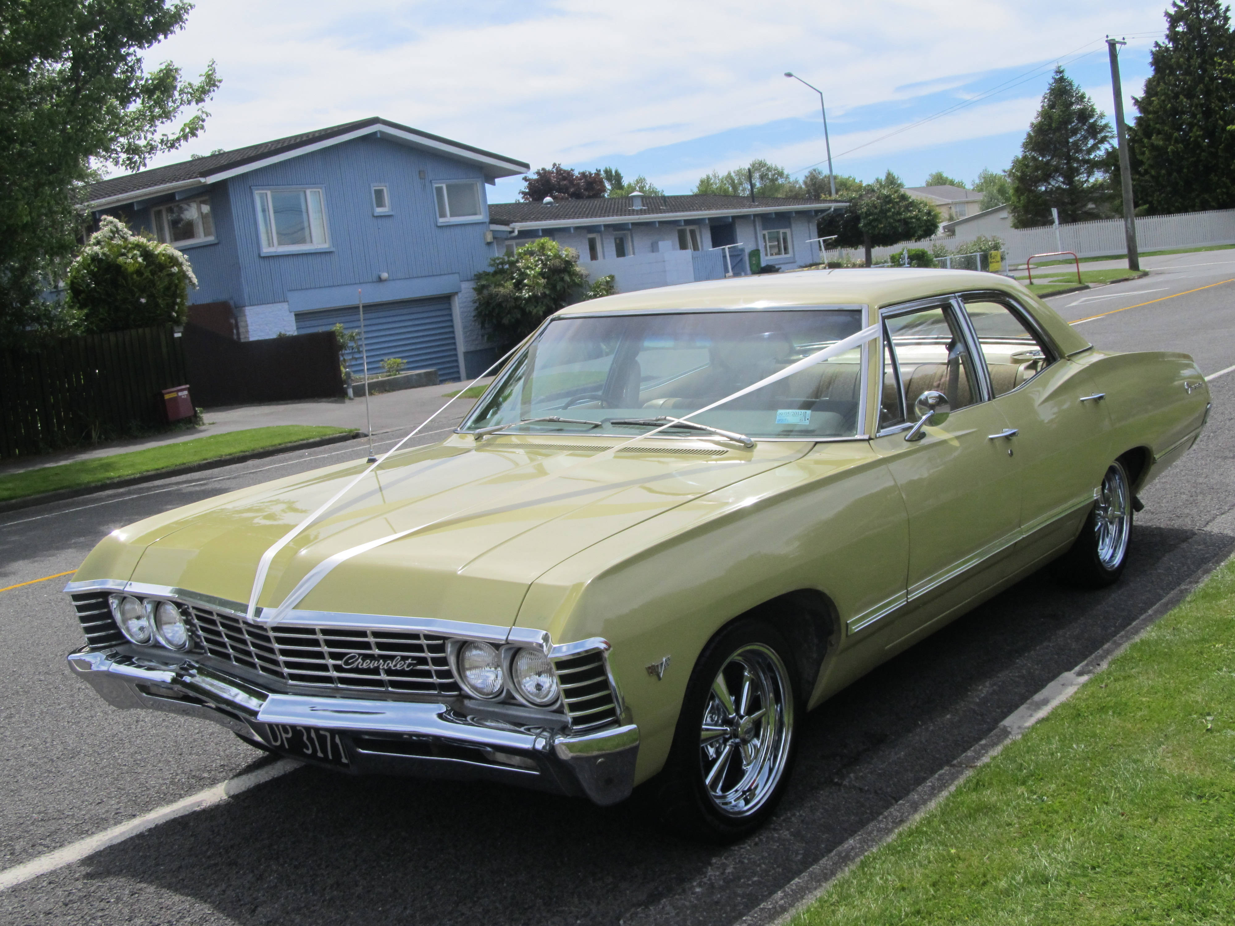 chevrolet impala 1967 桌布 壁紙 — 一輛復古橄欖綠色的雪佛蘭轎車，帶有鍍鉻細節，停在郊區街道上，背景是明亮的藍天。