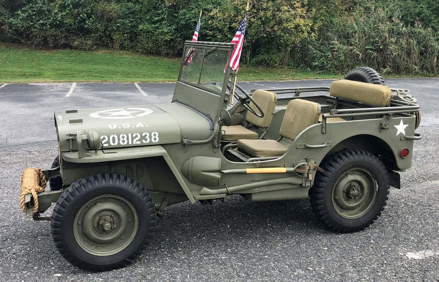 Jeep Willys 桌布 壁紙 — 一輛復古橄欖綠軍用吉普車，帶有美國國旗和星形徽章，停在碎石上，靠近綠色樹葉。