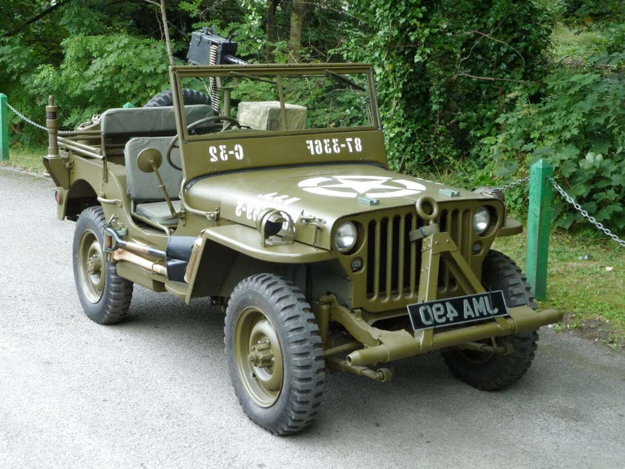 Jeep Willys 桌布 壁紙 — 一輛復古橄欖綠軍用吉普車，裝有機槍，停泊在碎石路上，周圍環繞著茂密的綠色植物。