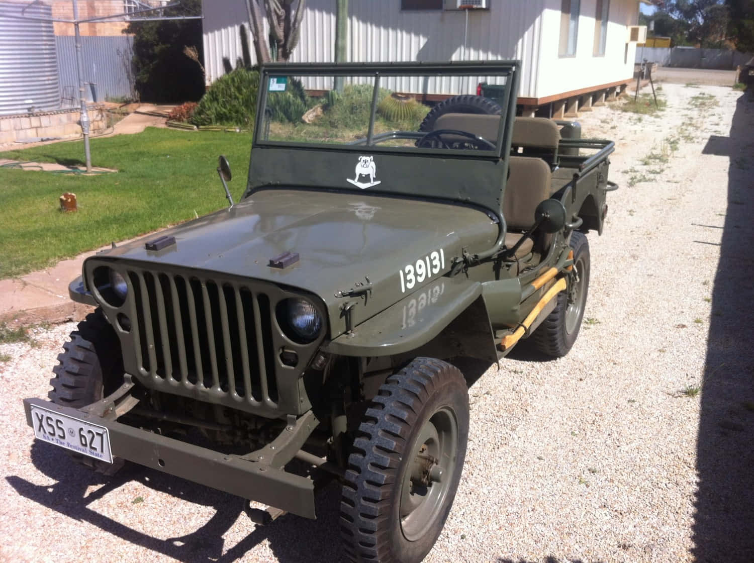 Jeep Willys 桌布 壁紙 — 一輛復古橄欖綠軍用吉普車停在白色房屋前的碎石路上，周圍有草地和樹木，陽光普照。