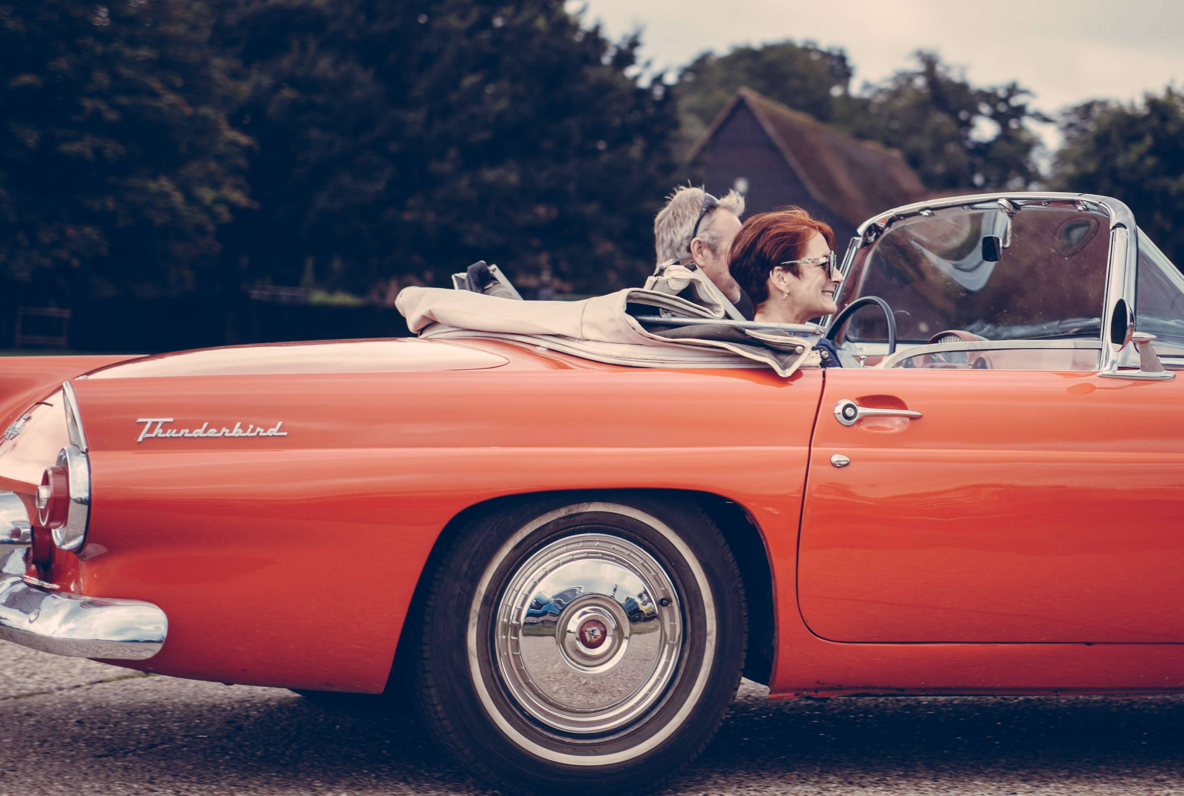 Love Story 桌布 壁紙 — 一輛復古橙色 Thunderbird 敞篷車沿著綠樹成蔭的道路行駛，一對微笑的夫婦享受著露天駕駛的樂趣。