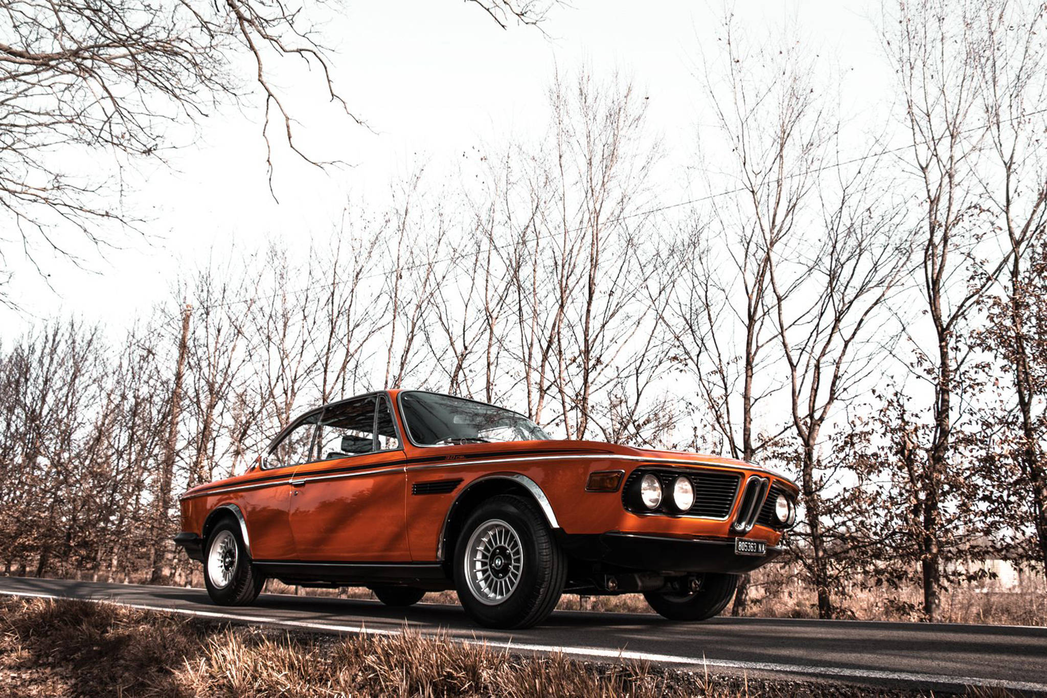 classic bmw 桌布 壁紙 — 一輛復古橙色BMW敞篷跑車停泊在寧靜的道路上，兩旁是光禿禿的冬季樹木，天空蒼白。