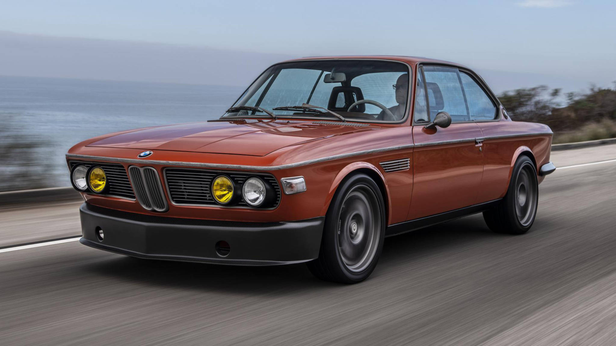 classic bmw 桌布 壁紙 — 一輛復古橙色BMW雙門跑車沿著海岸公路疾駛，背景是模糊的海洋和天空。