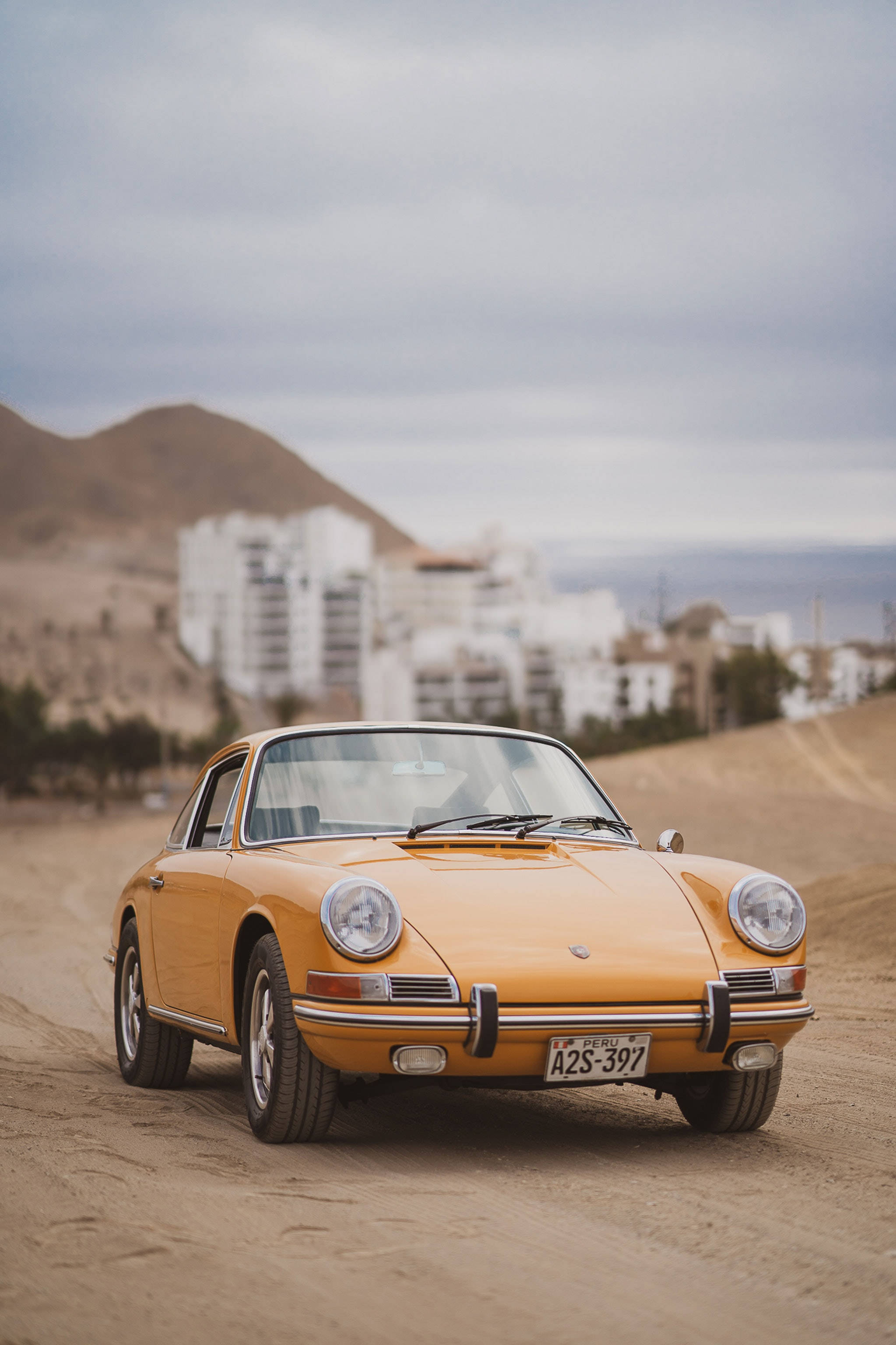 Retro 桌布 壁紙 — 一輛復古橙色保時捷911在塵土飛揚的沙漠公路上行駛，背景是白色建築物和山丘，天空多雲。