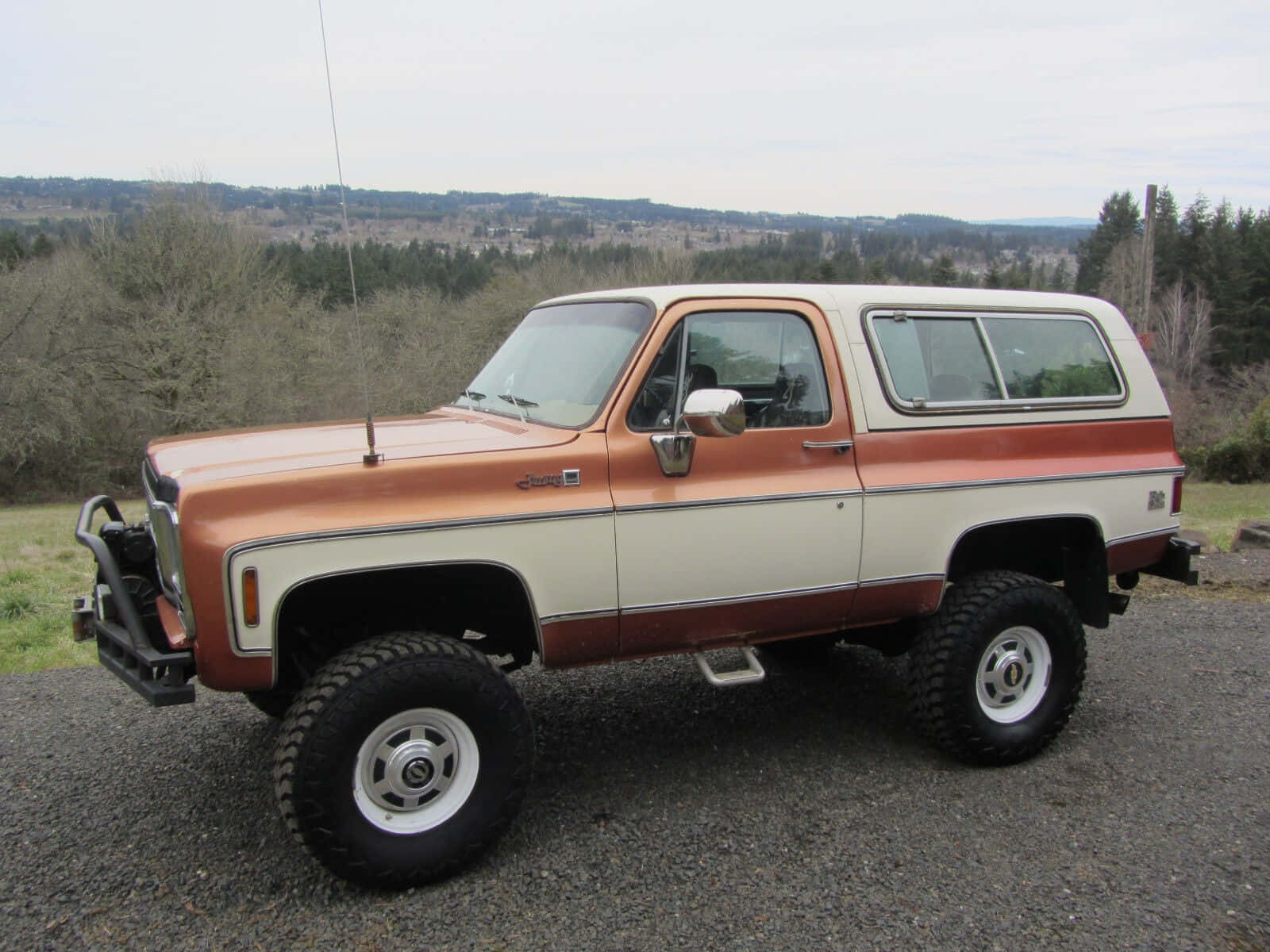 Gmc Jimmy 桌布 壁紙 — 一輛復古橙色和奶油色升高底盤的雪佛蘭 Blazer SUV 停在碎石路上，背景是森林覆蓋的山坡。