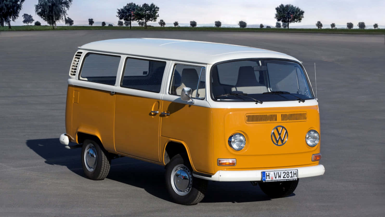 Van 桌布 壁紙 — 一輛復古橙色和白色的福斯 Volkswagen Type 2 廂型車停放在廣闊的灰色瀝青場地上，天空晴朗，遠處有稀疏的樹木。