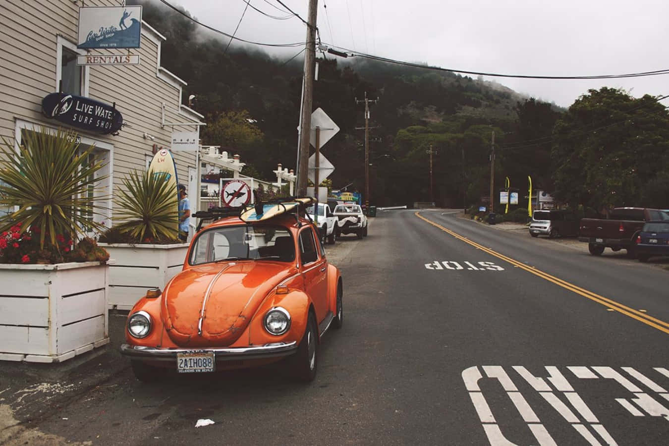 Vintage Surf 桌布 壁紙 — 一輛復古橙色福斯金龜車，車頂載著衝浪板，停泊在陰天下海邊道路旁的一間衝浪店旁邊。