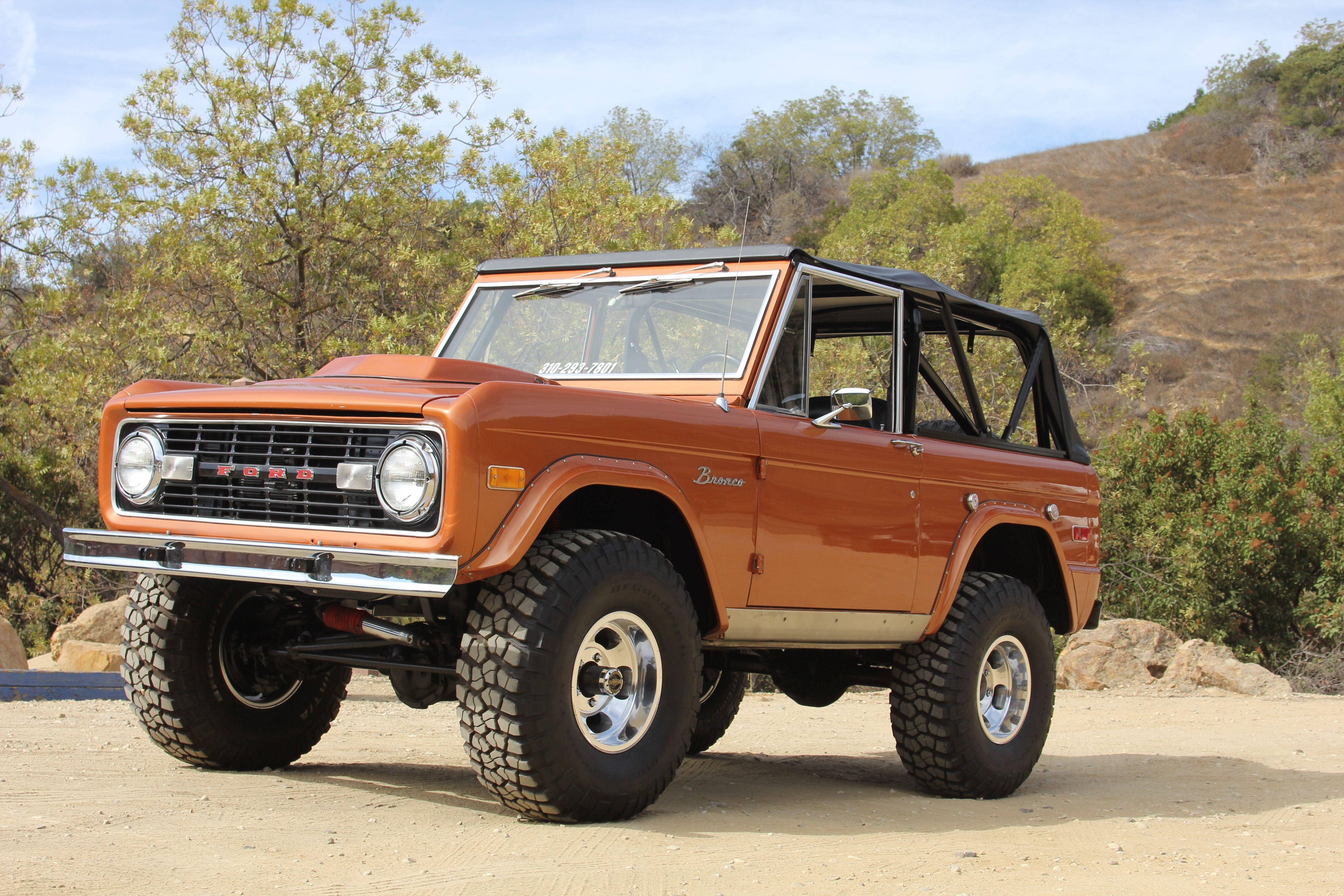 Ford Bronco 桌布 壁紙 — 一輛復古橙色福特Bronco，帶黑色防滾架，停在塵土飛揚的地形上，天空清澈湛藍。