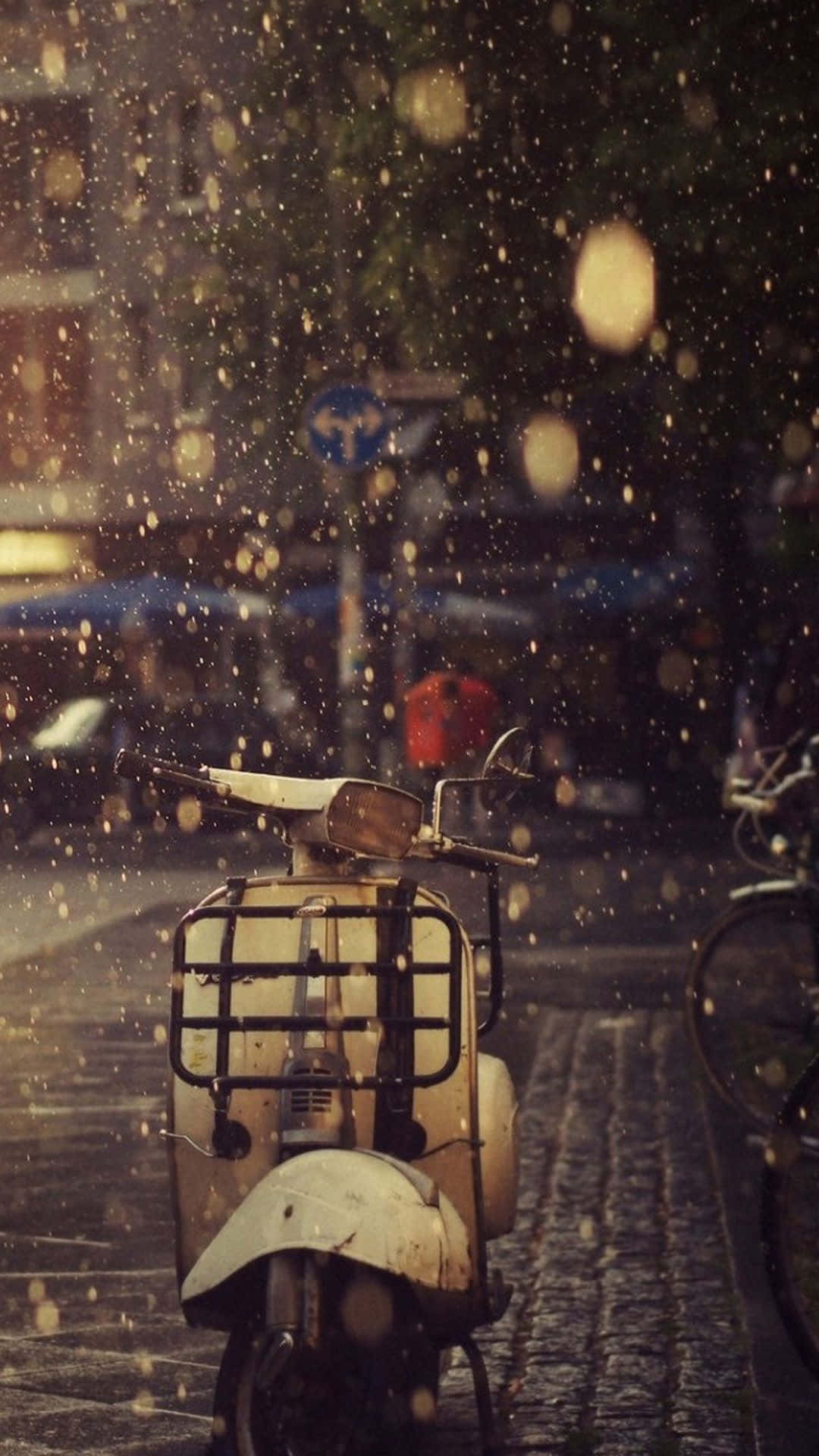 scooter 桌布 壁紙 — 一輛復古機車停在濕漉漉的鵝卵石街道上，金色的雨滴將城市背景模糊成柔和散景。