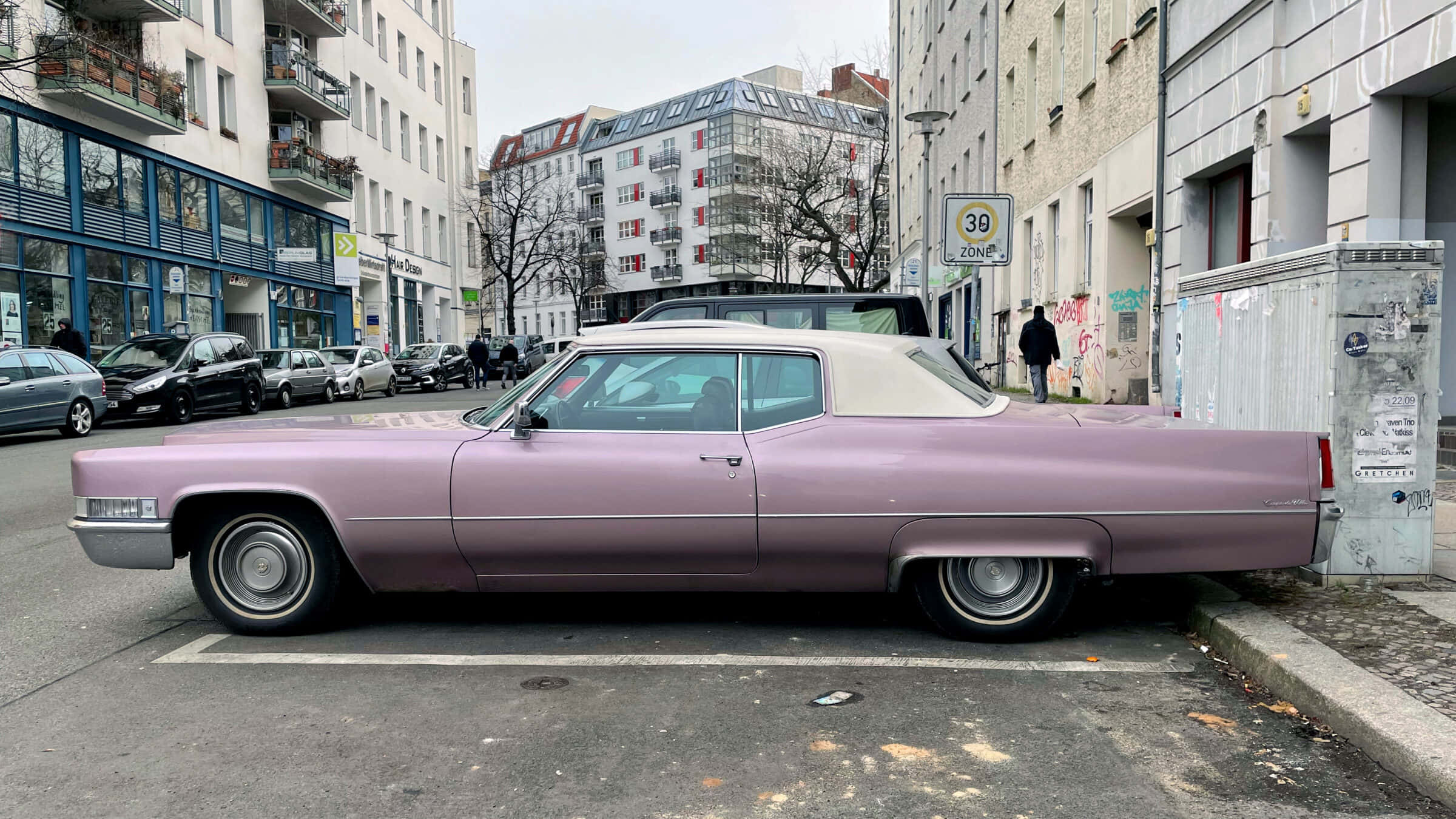 Pink Cadillac 桌布 壁紙 — 一輛復古淡紫色凱迪拉克，白色車頂，停放在現代建築和光禿樹木林立的城市街道上。
