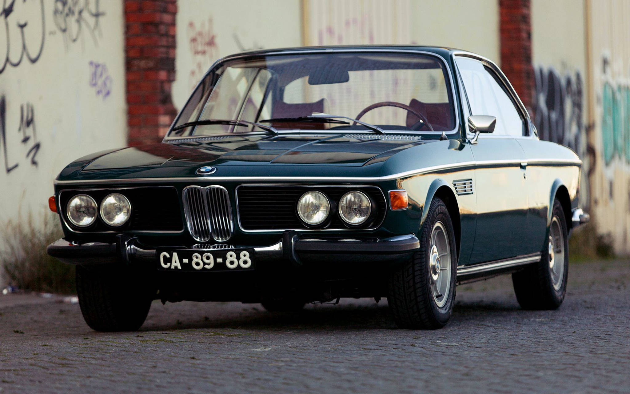 classic bmw 桌布 壁紙 — 一輛復古深綠色寶馬6系轎跑車停泊在鵝卵石街道上，旁邊是佈滿塗鴉的牆壁。