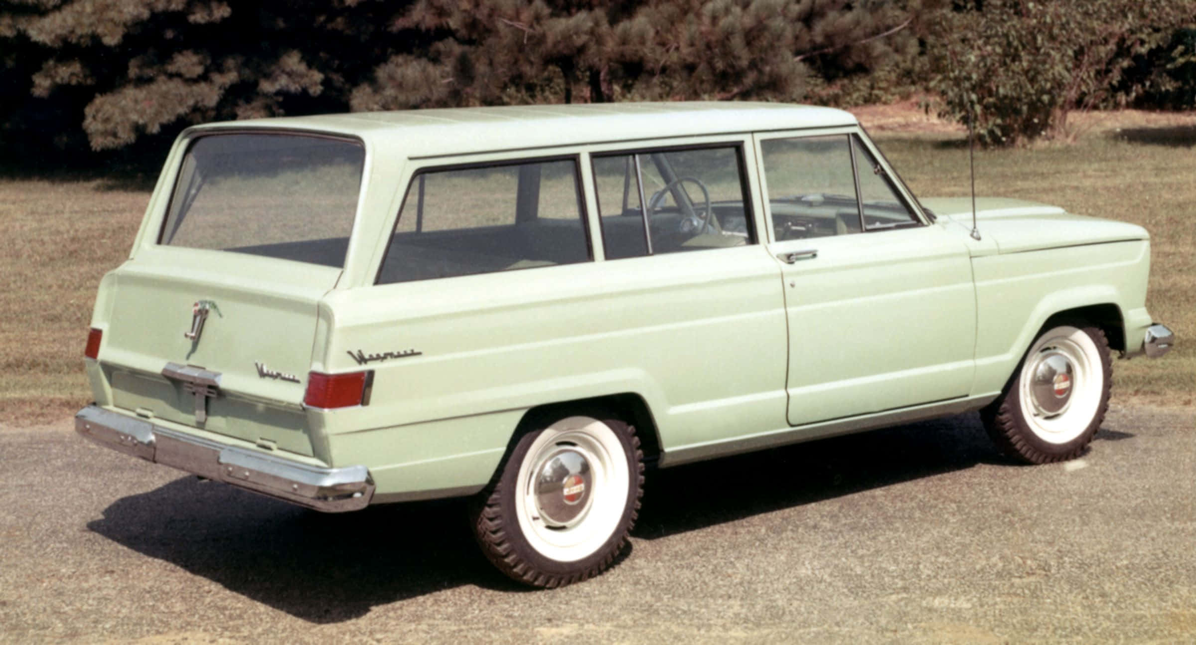 Jeep Wagoneer 桌布 壁紙 — 一輛復古淺綠色旅行車停在碎石上，背景有樹木，光線明亮。