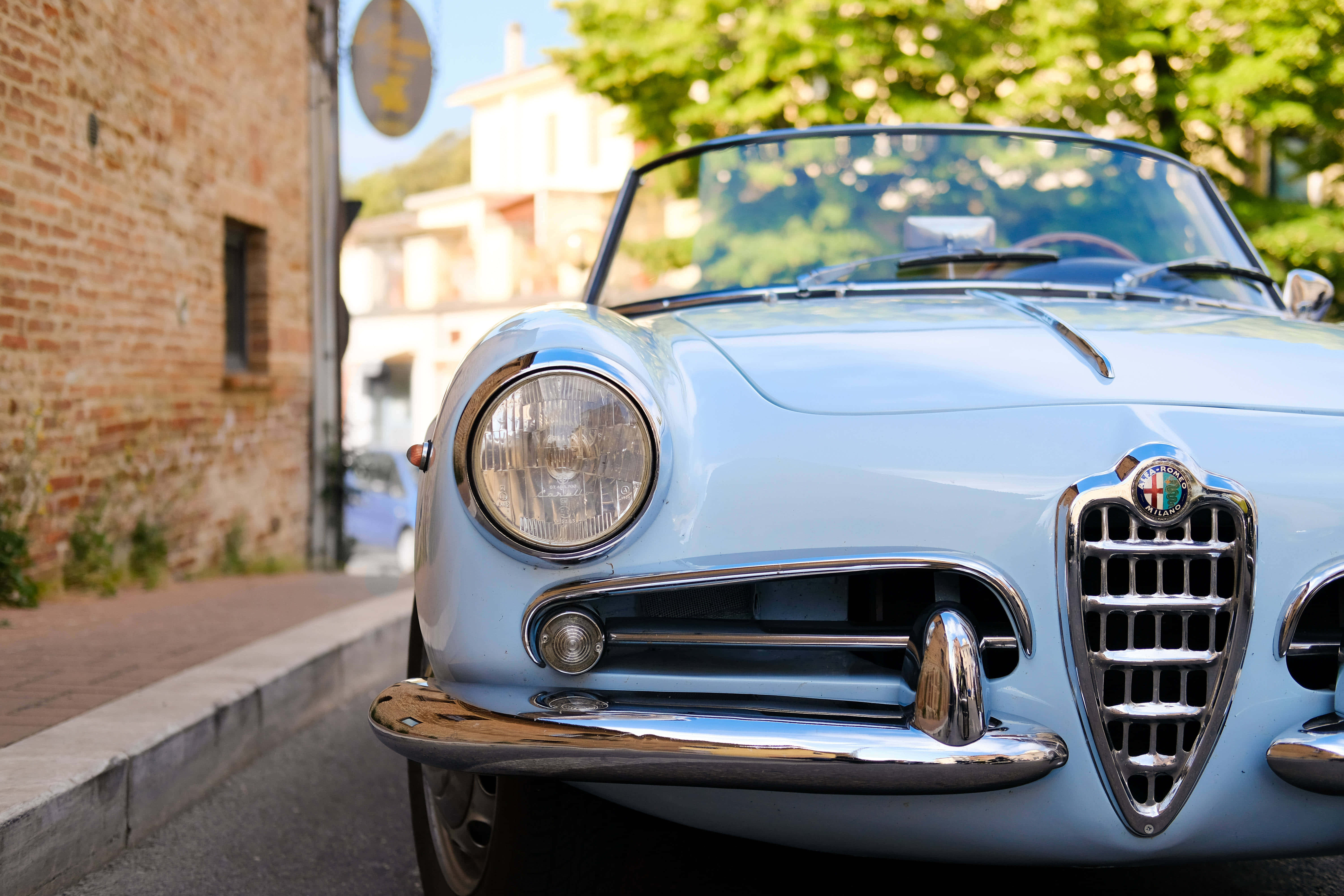 Alfa Romeo Spider 桌布 壁紙 — 一輛復古淺藍色愛快羅密歐蜘蛛跑車停泊在陽光普照的鵝卵石街道上，旁邊是磚牆和綠樹。