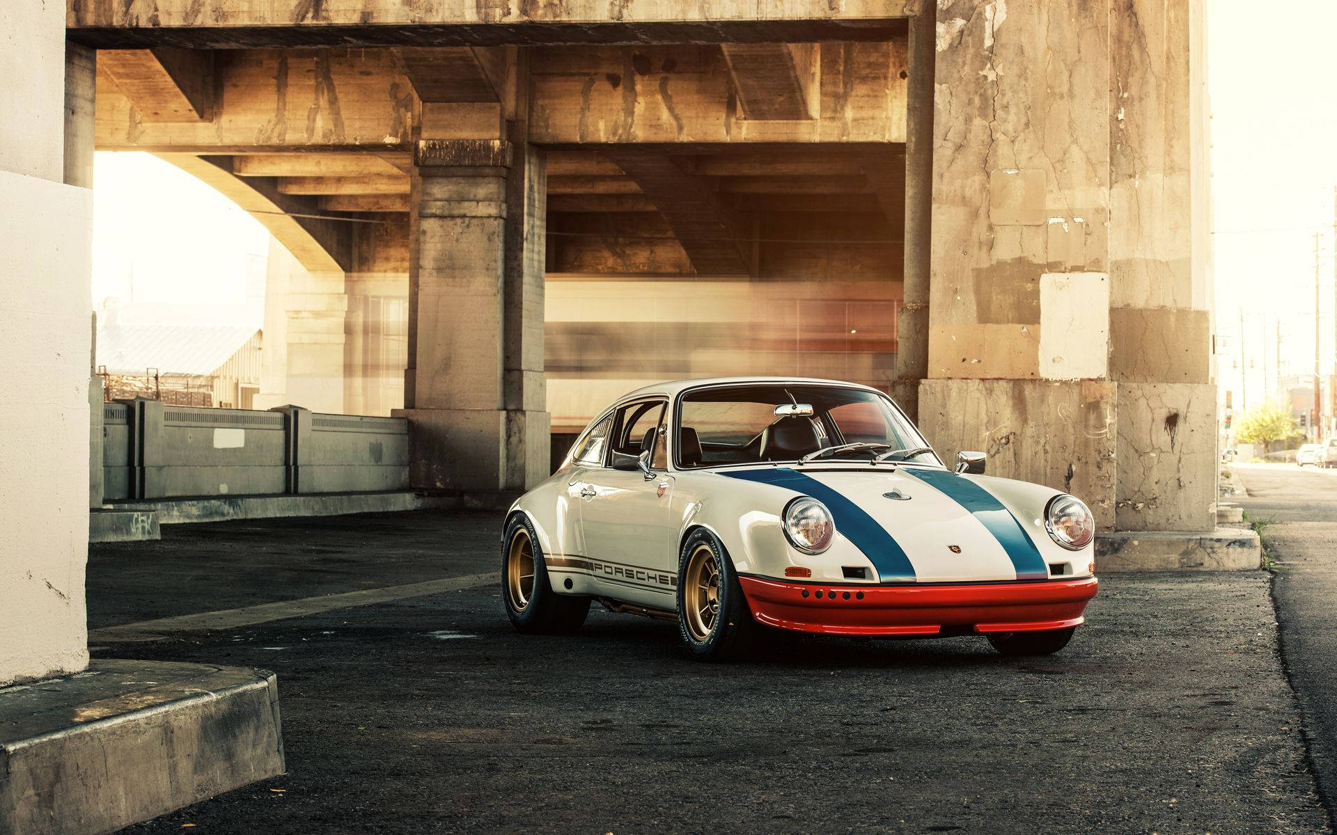 Porsche 911 桌布 壁紙 — 一輛復古白色保時捷911，帶有藍色和紅色賽車條紋，停在風化的混凝土天橋下，沐浴在金色時刻的光線中。