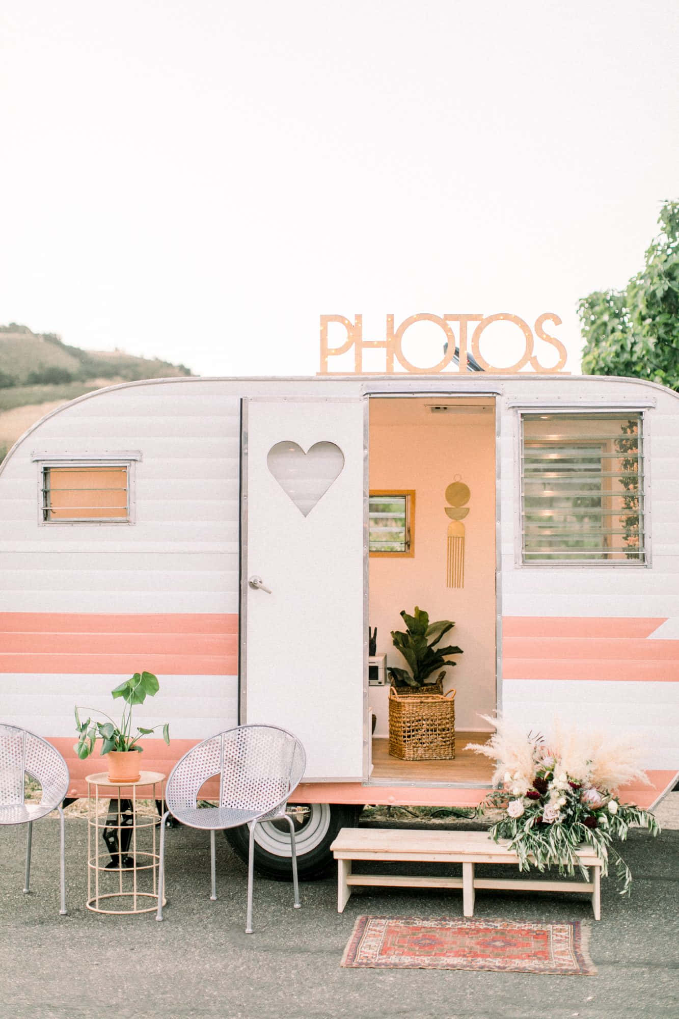 Photo Booth 桌布 壁紙 — 一輛復古白色和珊瑚色的露營車，帶有心形門和「PHOTOS」標誌，提供迷人的戶外拍照亭，配有椅子和花卉裝飾。