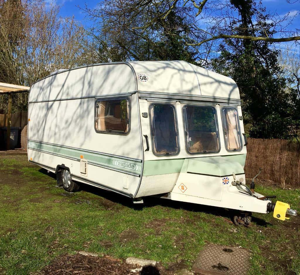Caravan 桌布 壁紙 — 一輛復古白色和綠色的露營車在明亮的藍天下休息在草地上，光禿禿的樹木投下陰影。