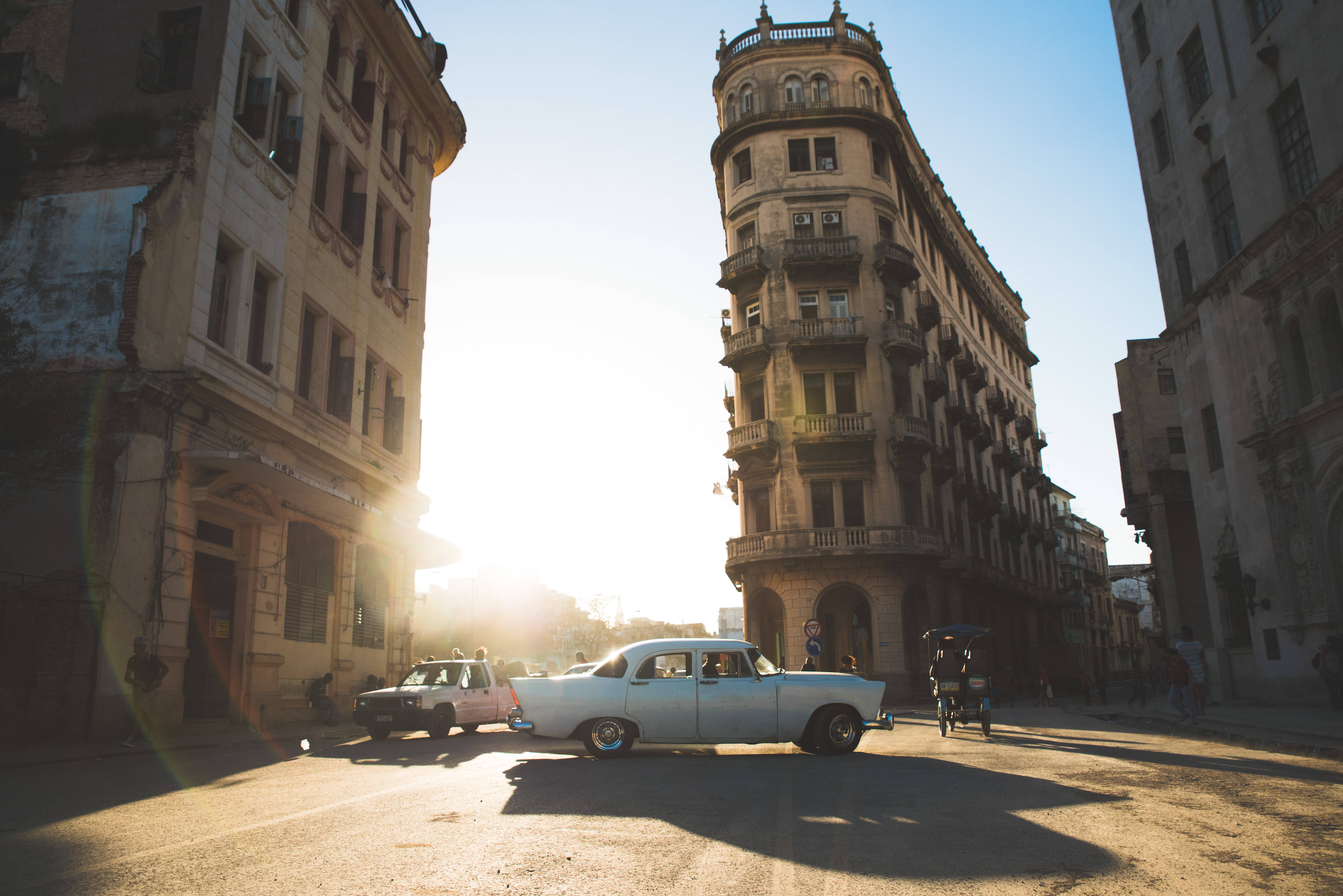 Cuba 桌布 壁紙 — 一輛復古白色汽車停泊在陽光普照的街道上，兩旁是飽經風霜的殖民時期建築，溫暖的金色陽光投下長長的影子。