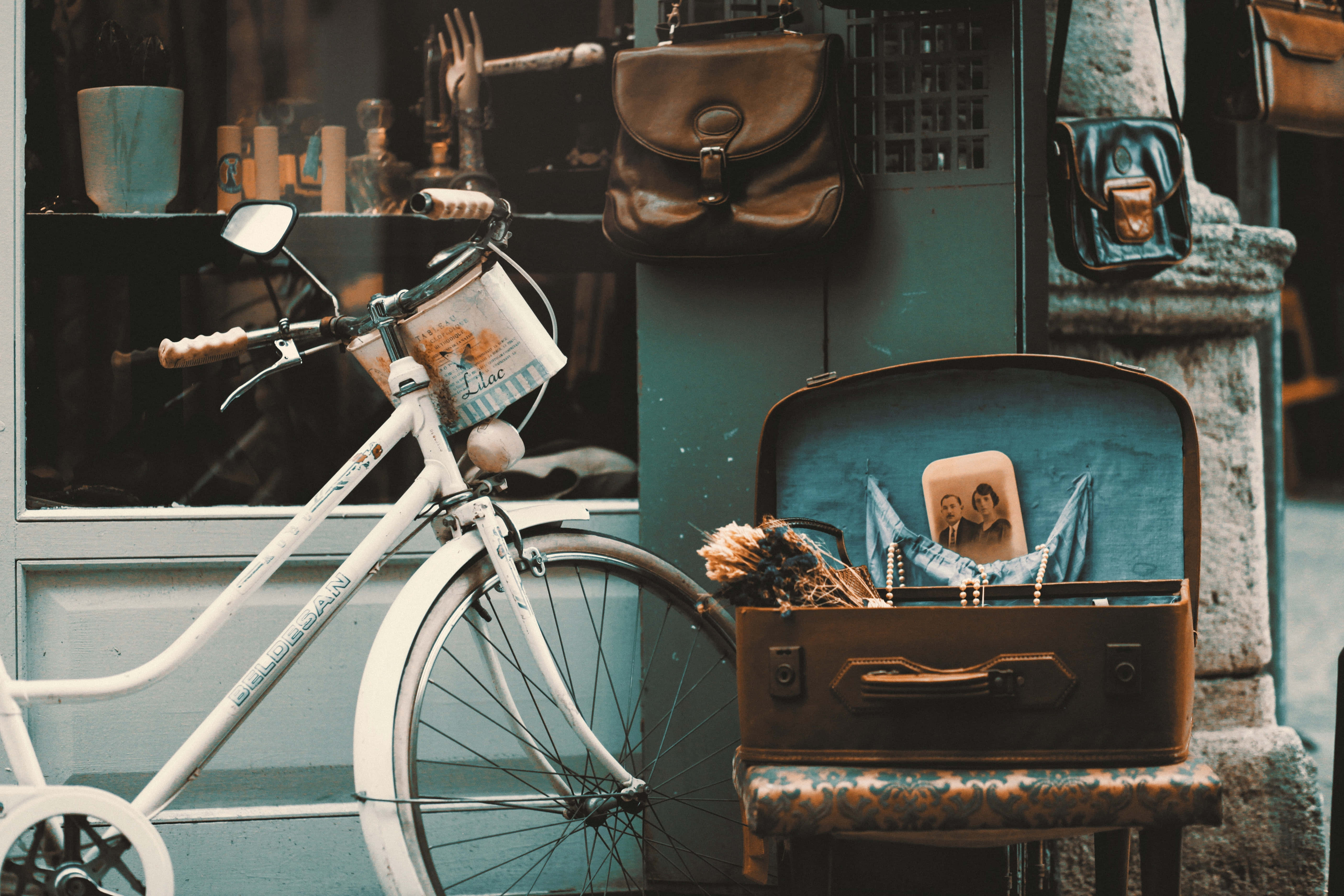 Cute Vintage 桌布 壁紙 — 一輛復古白色腳踏車靠在一旁，旁邊有一個打開的行李箱，裡面放著一張照片和乾燥花，行李箱放在店外一張有圖案的凳子上。