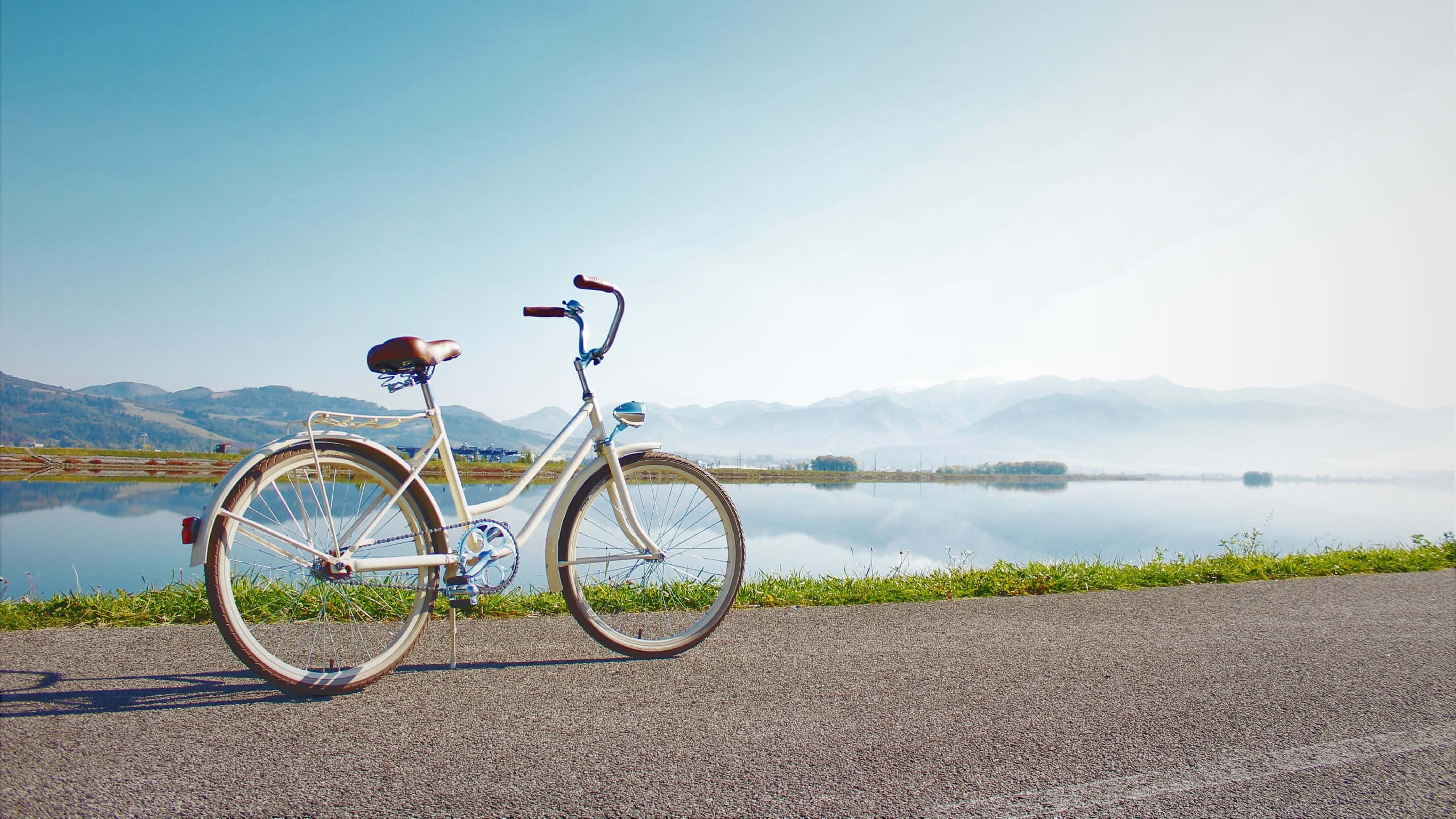 Bicycle 桌布 壁紙 — 一輛復古白色自行車停放在平靜湖泊旁的鋪砌道路上，背景是霧氣繚繞的山脈和清澈的藍天。