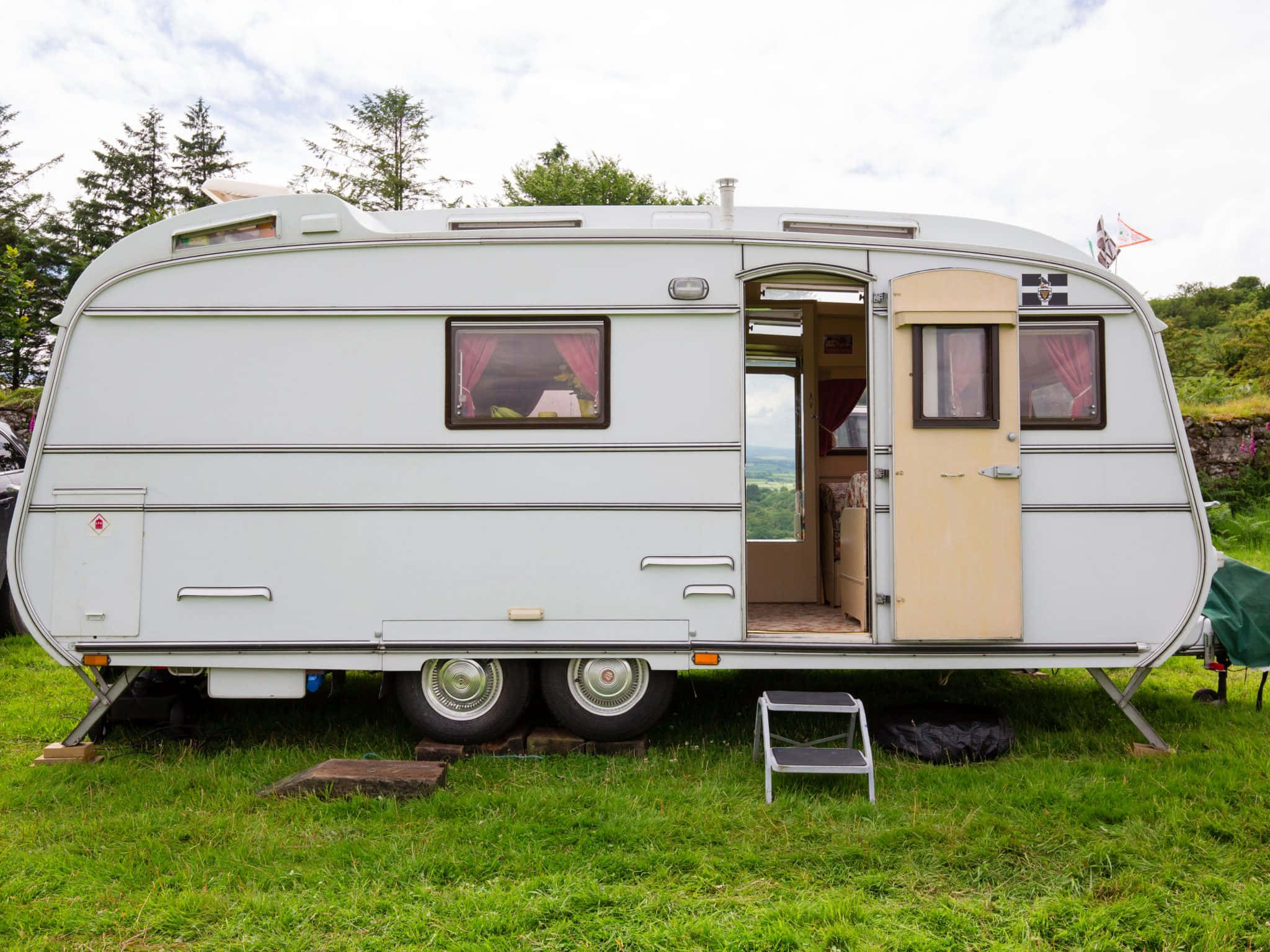 Caravan 桌布 壁紙 — 一輛復古白色露營拖車打開車門，停在草地上，背景有樹木和山丘。