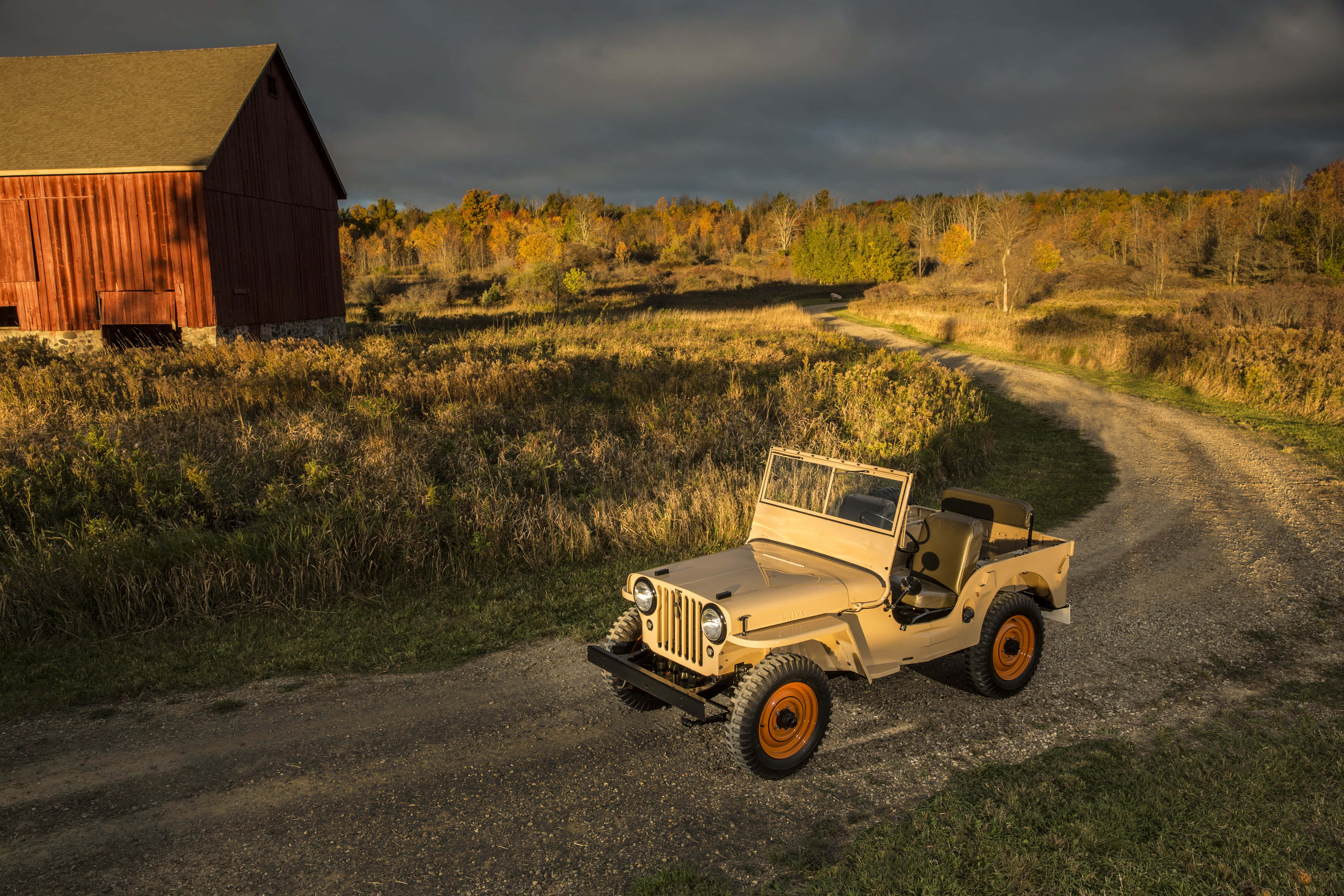 Jeep Cj 桌布 壁紙 — 一輛復古米色吉普車配橙色車輪，停在碎石路上，靠近紅色穀倉，背景是戲劇性的秋日天空。
