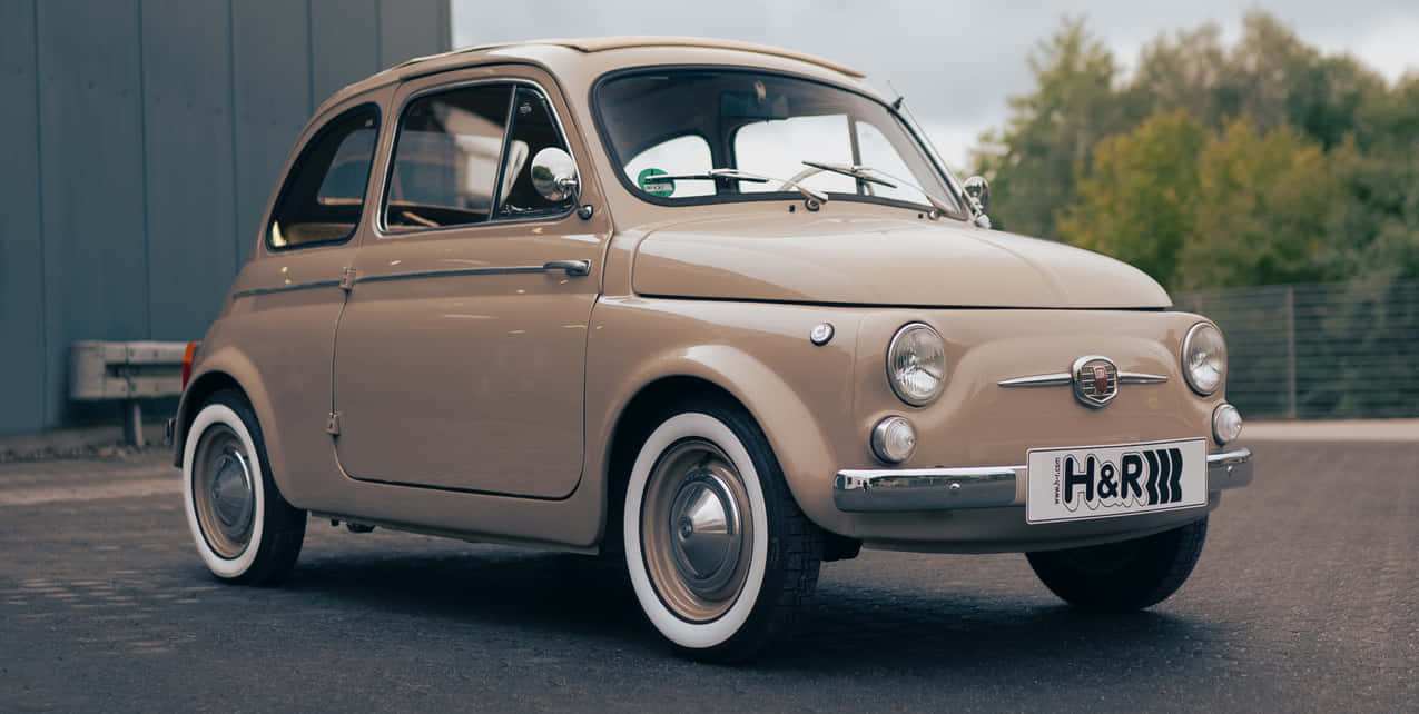 Fiat 桌布 壁紙 — 一輛復古米色菲亞特500停在瀝青路上，背景有樹木和灰色建築物。
