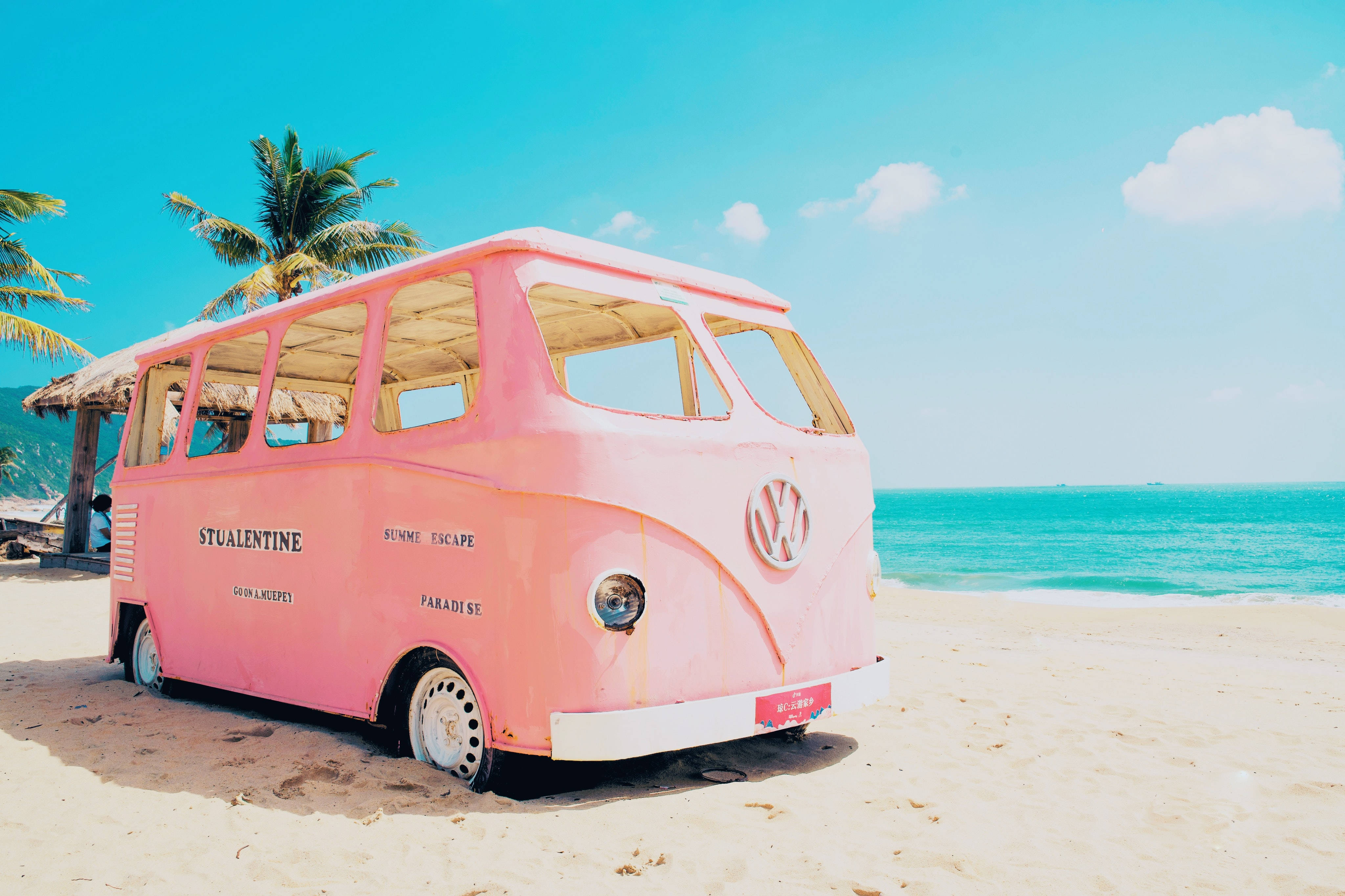 Beach Desktop 桌布 壁紙 — 一輛復古粉紅色福斯露營車在陽光明媚的海灘上休息，旁邊是碧綠的海水和棕櫚樹。