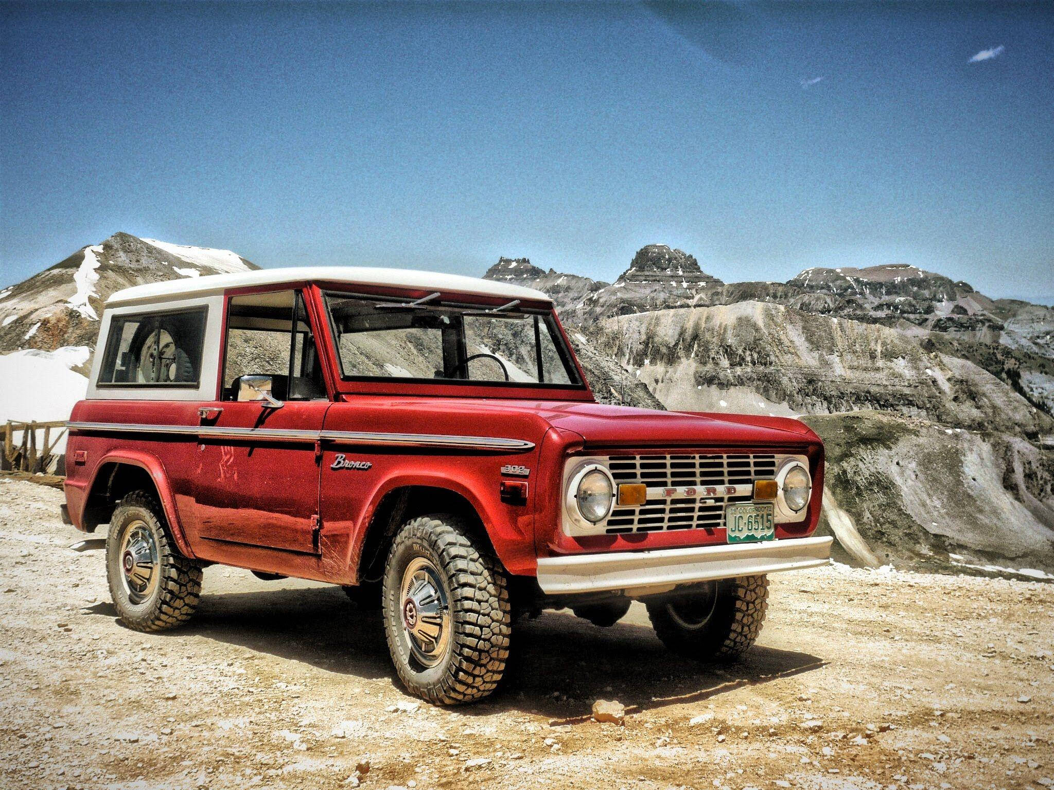 Ford Bronco 桌布 壁紙 — 一輛復古紅色Bronco SUV停泊在白雪覆蓋的山路上，背景是晴朗的藍天和崎嶇的山峰。