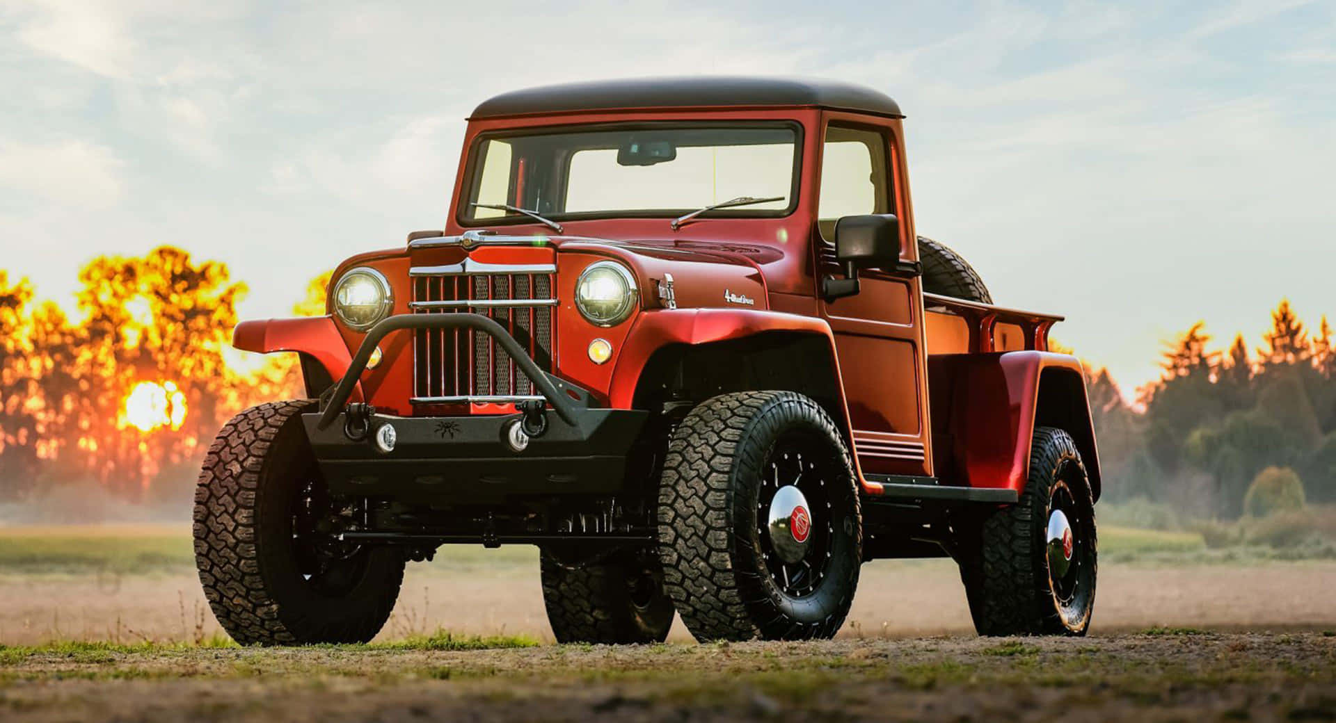 Jeep Willys 桌布 壁紙 — 一輛復古紅色Jeep Willys，黑色車頂和超大輪胎，停在日落時分的泥地上，背景是金色的樹木。