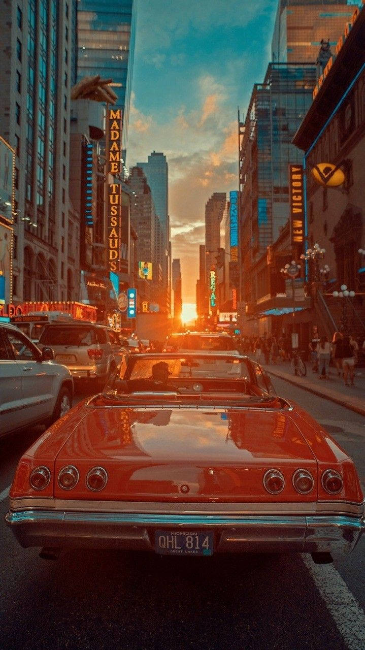 Vintage Car 桌布 壁紙 — 一輛復古紅色敞篷車在日落時分行駛在繁華的城市街道上，霓虹燈在高樓大廈之間閃爍。