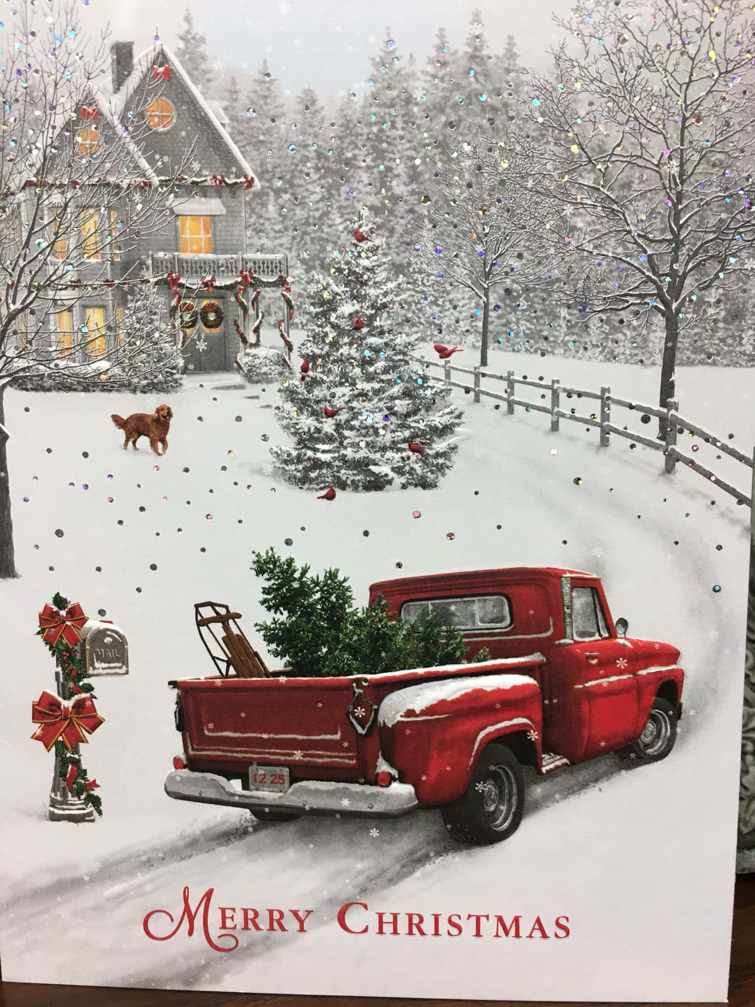 桌布 壁紙 — 一輛復古紅色皮卡車載著聖誕樹穿過白雪覆蓋的村莊街道，附近有一間溫暖亮燈的房子和一隻黃金獵犬。