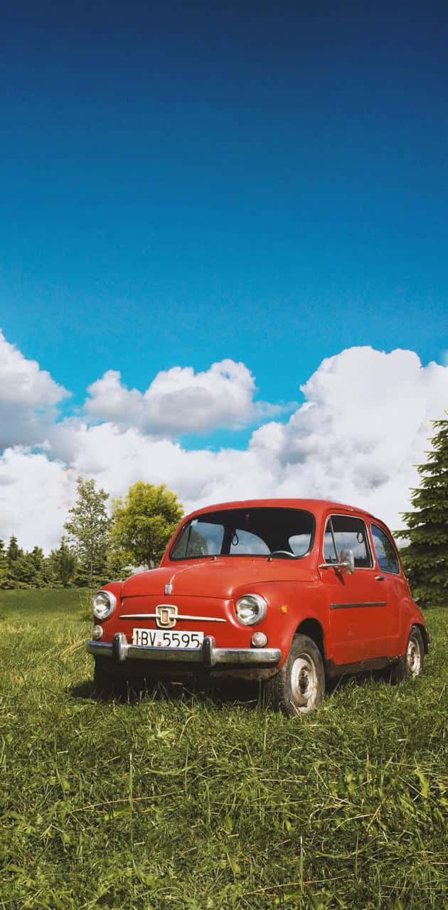 Vintage Car iPhone 桌布 壁紙 — 一輛復古紅色菲亞特500停泊在茂密的綠色田野中，天空湛藍，白雲朵朵。