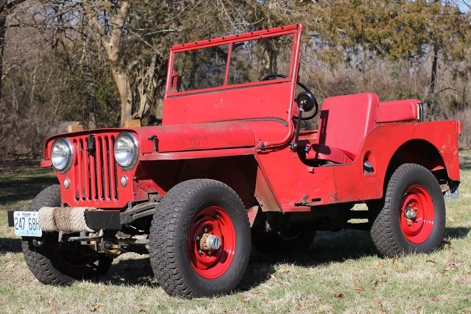 Jeep Willys 桌布 壁紙 — 一輛復古紅色軍用吉普車，配備堅固輪胎，停放在陽光普照的田野中、光禿禿的樹木下的草地上。