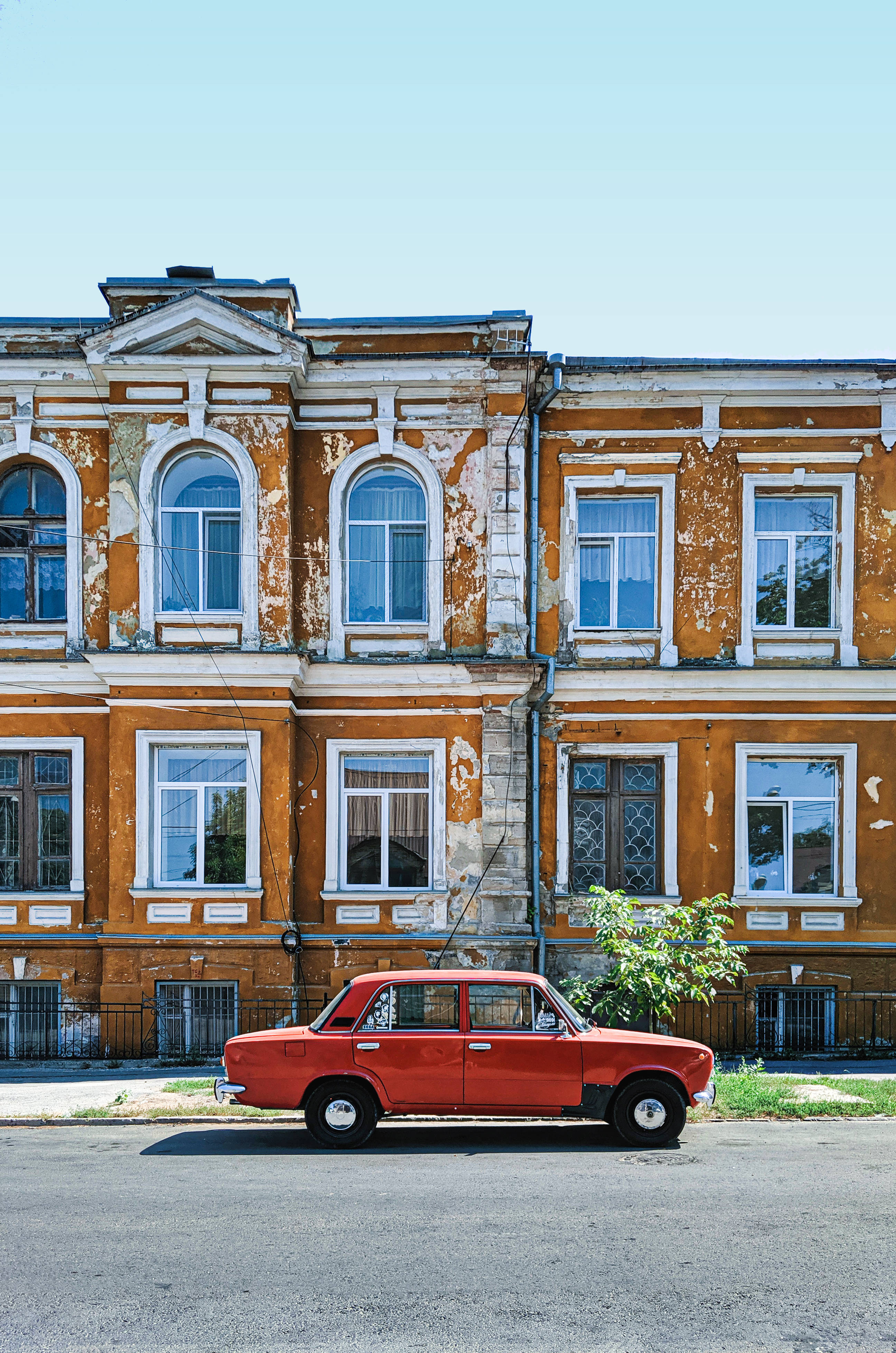 Moldova 桌布 壁紙 — 一輛復古紅色轎車停在一棟褪色的橙色建築前，建築物有剝落的油漆和拱形窗戶，背景是清澈的藍天。