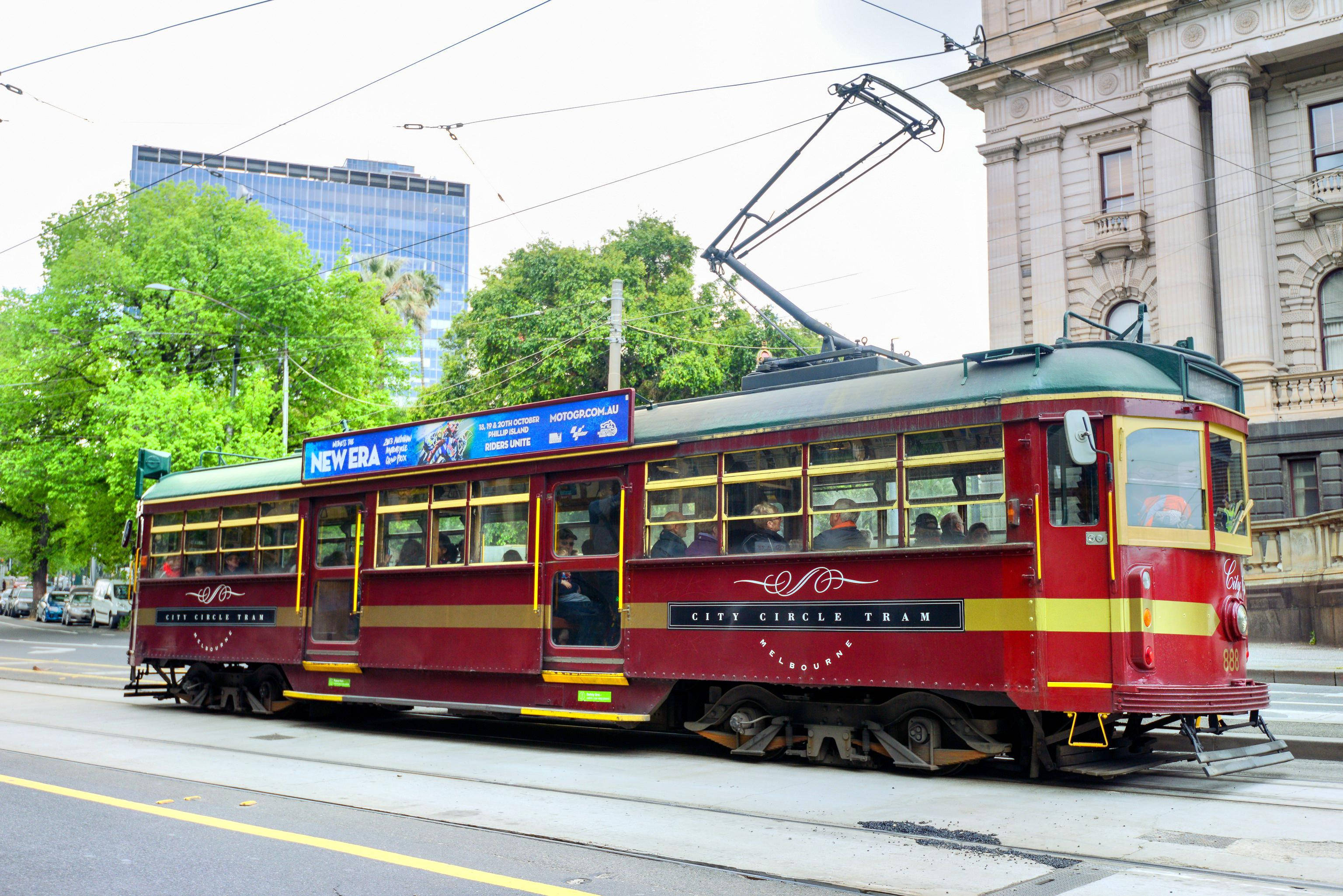 墨爾本 桌布 壁紙 — 一輛復古紅金色的墨爾本電車在陰天下行駛於綠樹成蔭、歷史建築林立的城市街道。