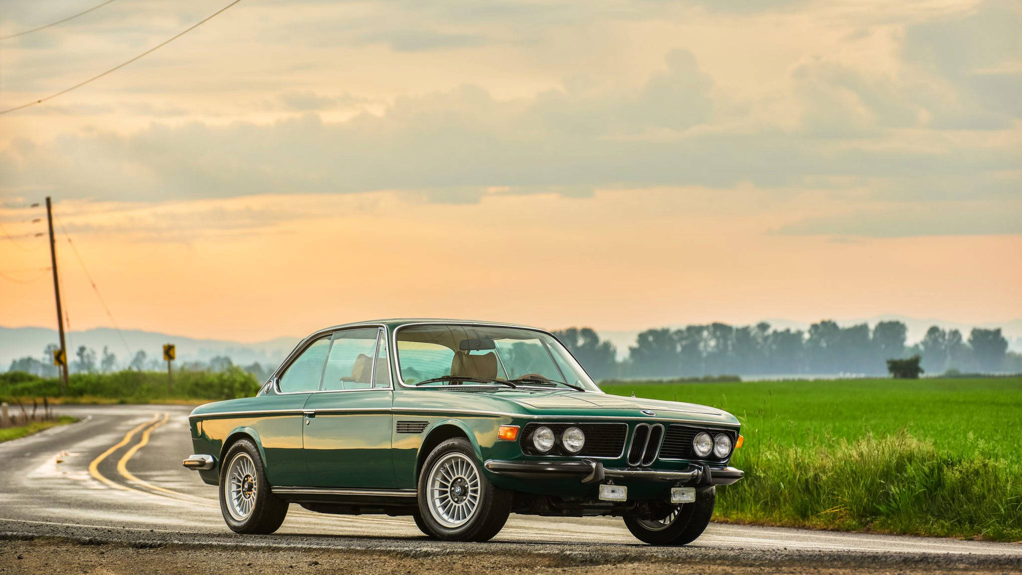 classic bmw 桌布 壁紙 — 一輛復古綠色BMW雙門跑車在日落時分行駛在鄉村道路上，伴隨著金色的雲彩和翠綠的田野。