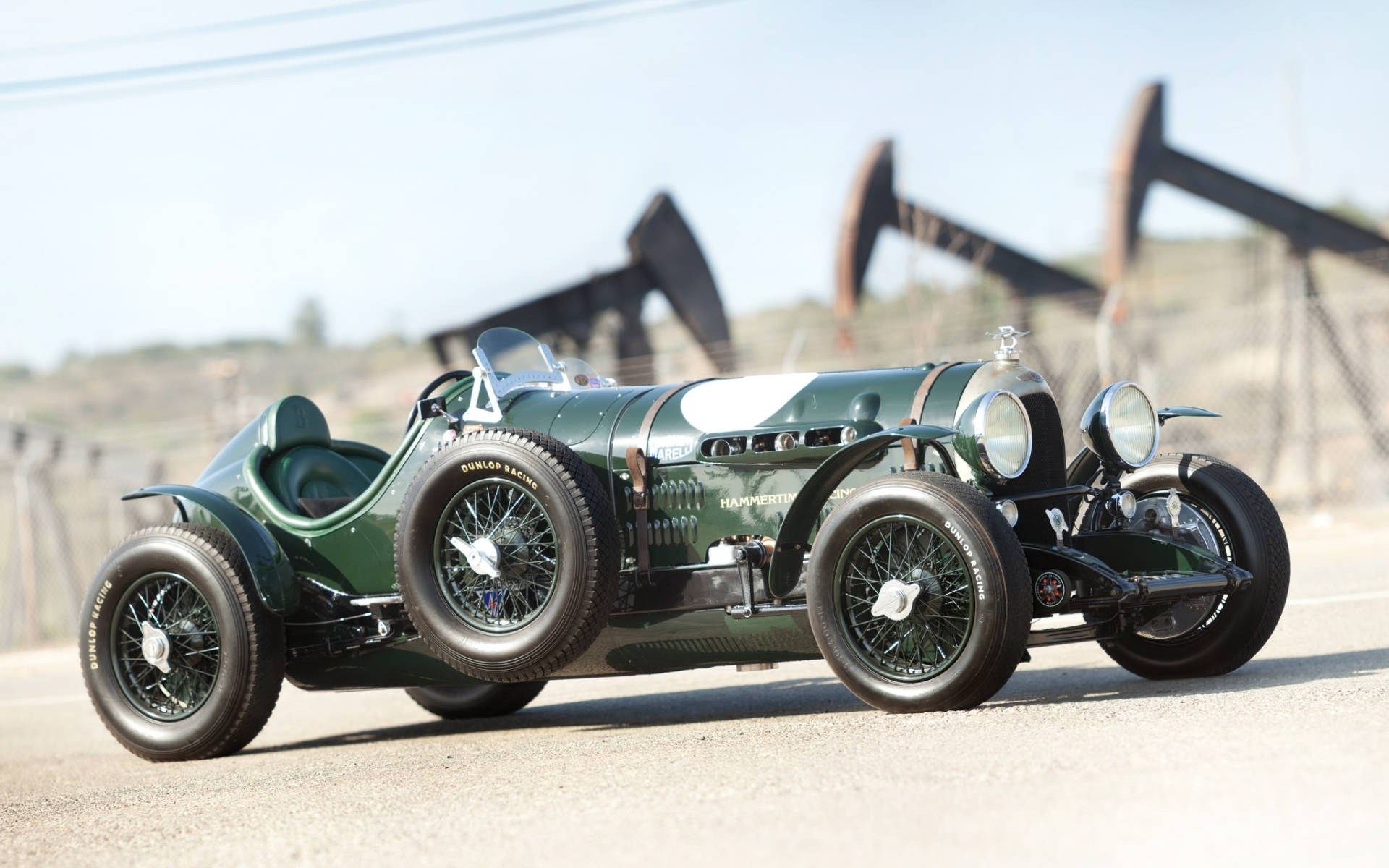bentley HD 桌布 壁紙 — 一輛復古綠色Hamer賽車，配備鋼絲輪圈，停放在鋪砌道路上，背景模糊的油泵在明亮日光下可見。
