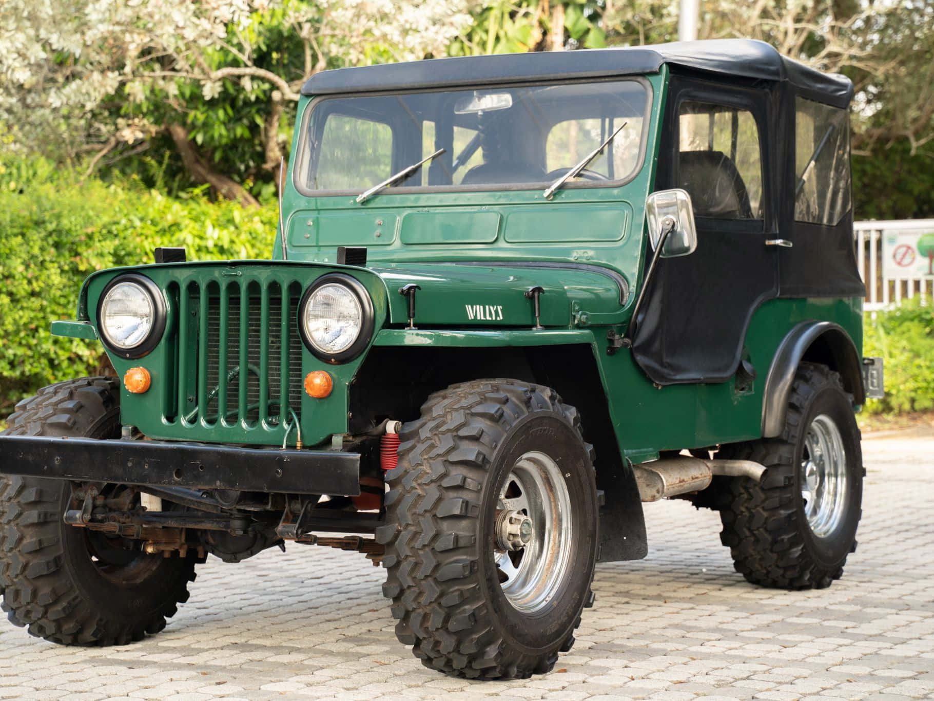 Jeep Willys 桌布 壁紙 — 一輛復古綠色Willys Jeep，配備黑色軟頂和堅固輪胎，停放在鋪砌的庭院中，四周環繞著茂密的綠植。
