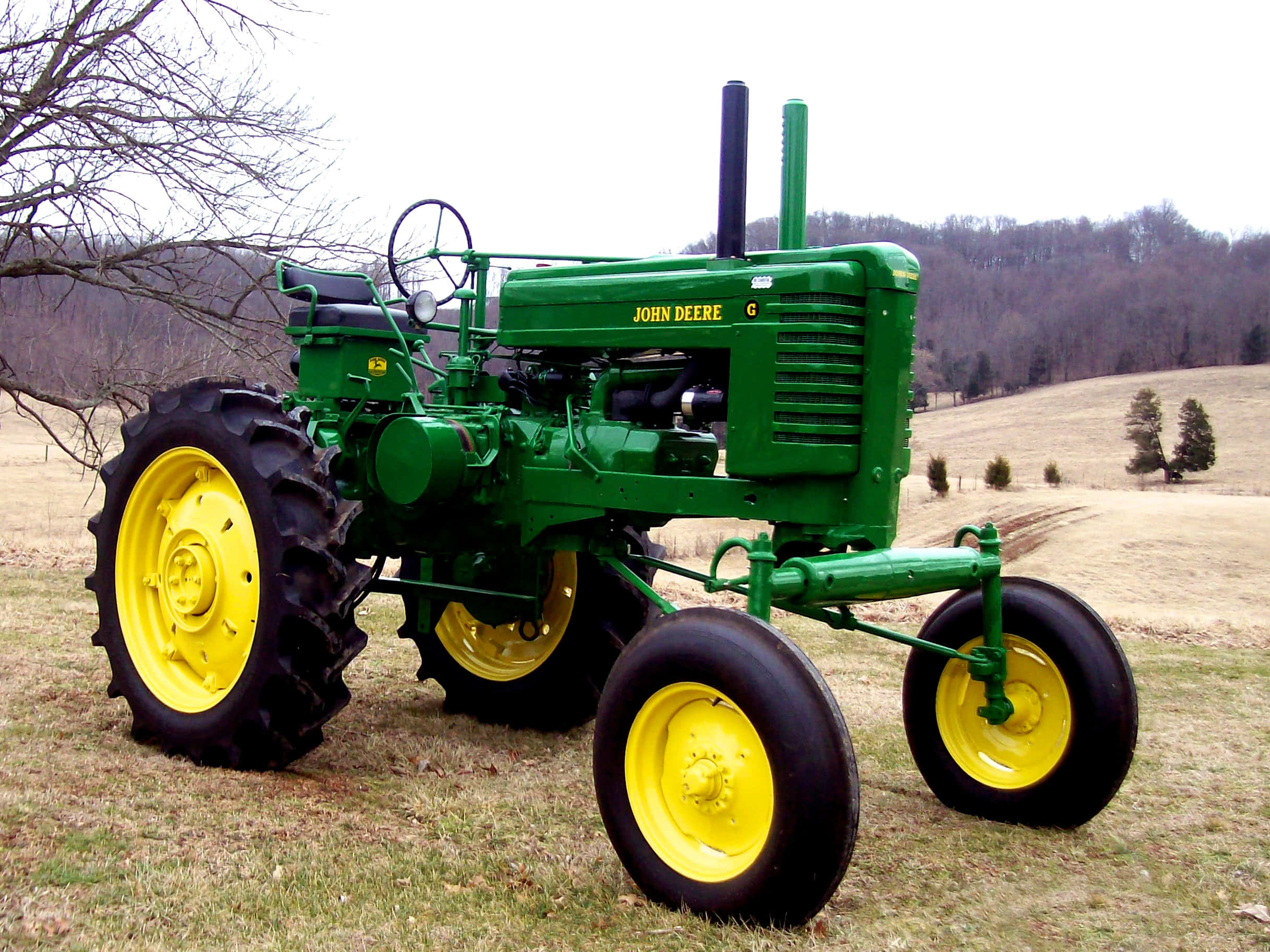 Tractor 桌布 壁紙 — 一輛復古綠色約翰迪爾拖拉機，帶有亮黃色車輪，站在草地上，背景是光禿禿的樹木和起伏的山丘，天空多雲。