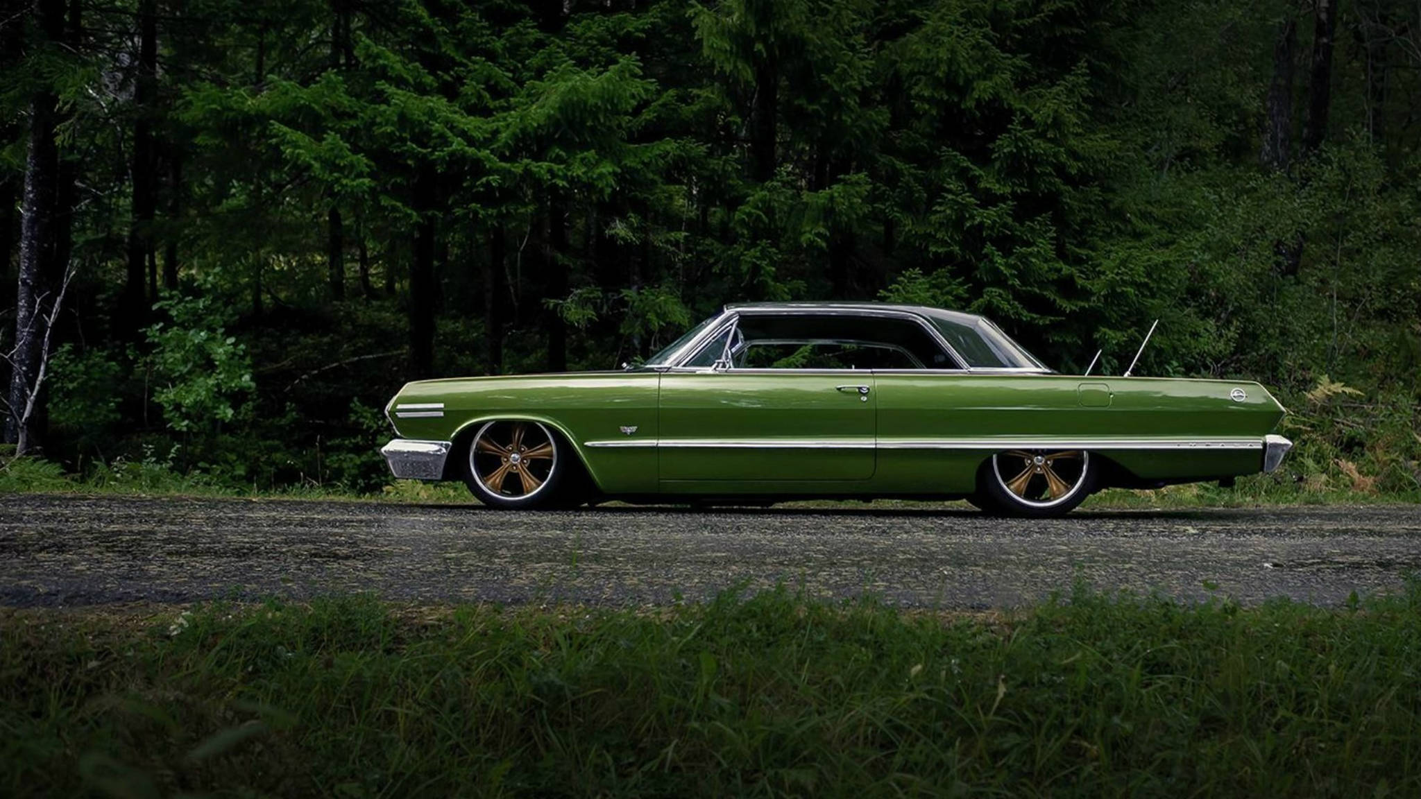 chevrolet impala 1967 桌布 壁紙 — 一輛復古綠色肌肉車配金色輪圈，停放在被茂密森林環繞的碎石路上。