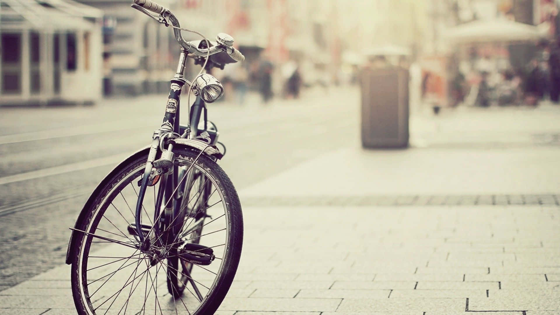 Bikes 桌布 壁紙 — 一輛復古腳踏車獨自停放在陽光普照的鵝卵石人行道上，背景是柔和模糊的城市街道。