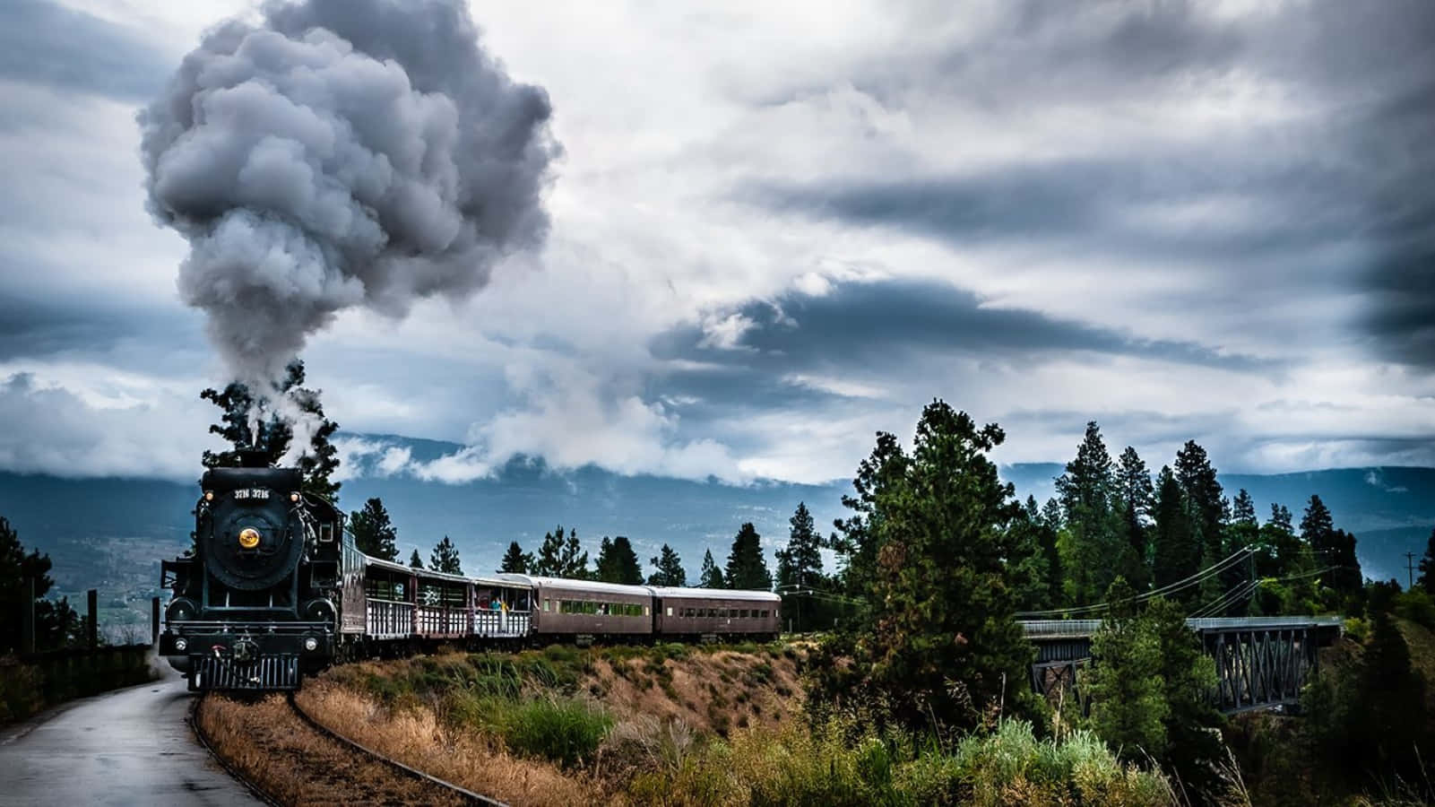 The Hogwarts Express Train 桌布 壁紙 — 一輛復古蒸汽火車在戲劇性的多雲天空下行駛穿過松樹林，噴出濃煙。