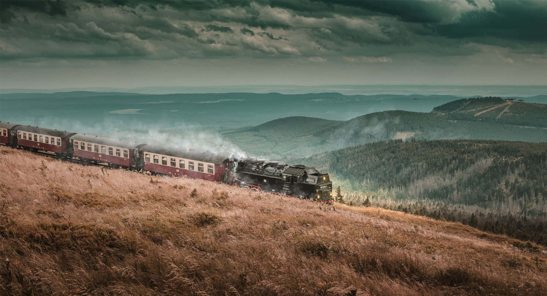 Hogwarts Express 桌布 壁紙 — 一輛復古蒸汽火車在暴風雨的天空下，沿著長滿青草的山坡行駛，遠處是連綿起伏的森林山丘。