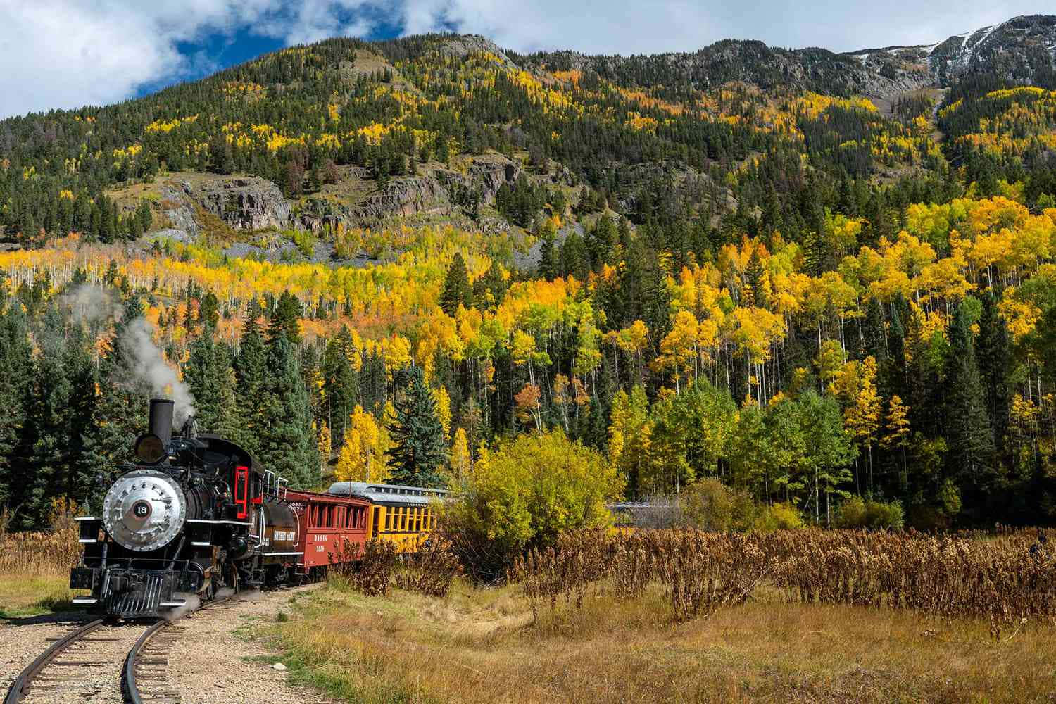 Colorado Mountain 桌布 壁紙 — 一輛復古蒸汽火車穿過充滿活力的秋日森林，金色白楊和常綠樹在岩石山脈前，天空部分多雲。