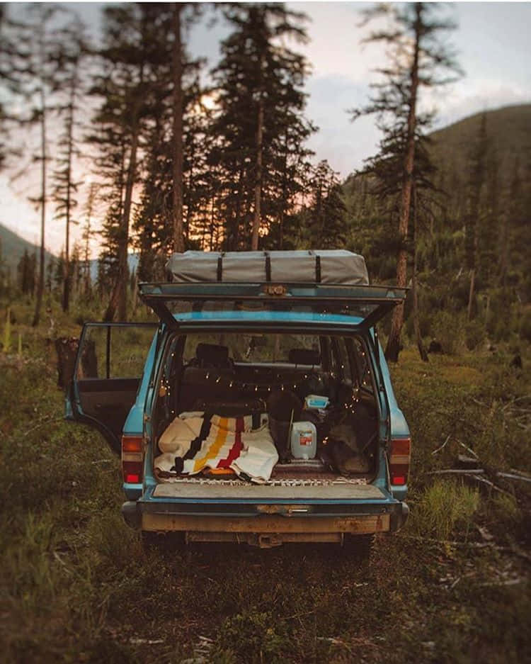 Minimalist Camping 桌布 壁紙 — 一輛復古藍色旅行車在日落時分停在森林中，後車廂打開，露出露營裝備和燈串。